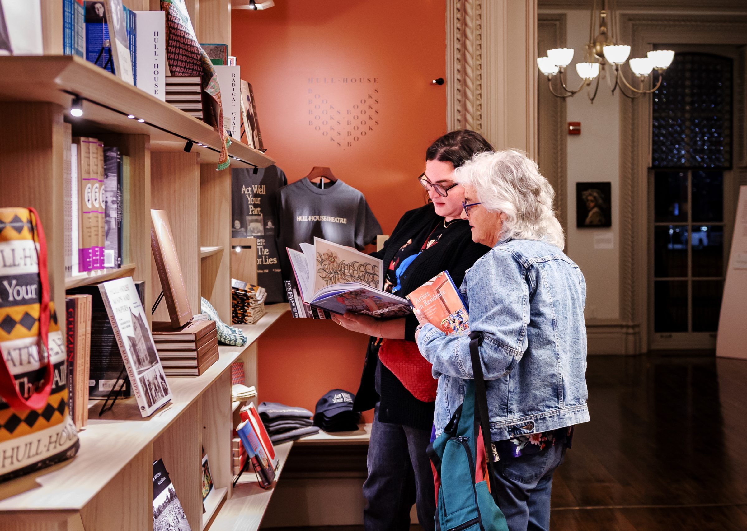 Two women stand in a museum gift shop, browsing books together near a display shelf filled with books and souvenirs. The room has ornate décor and warm lighting.