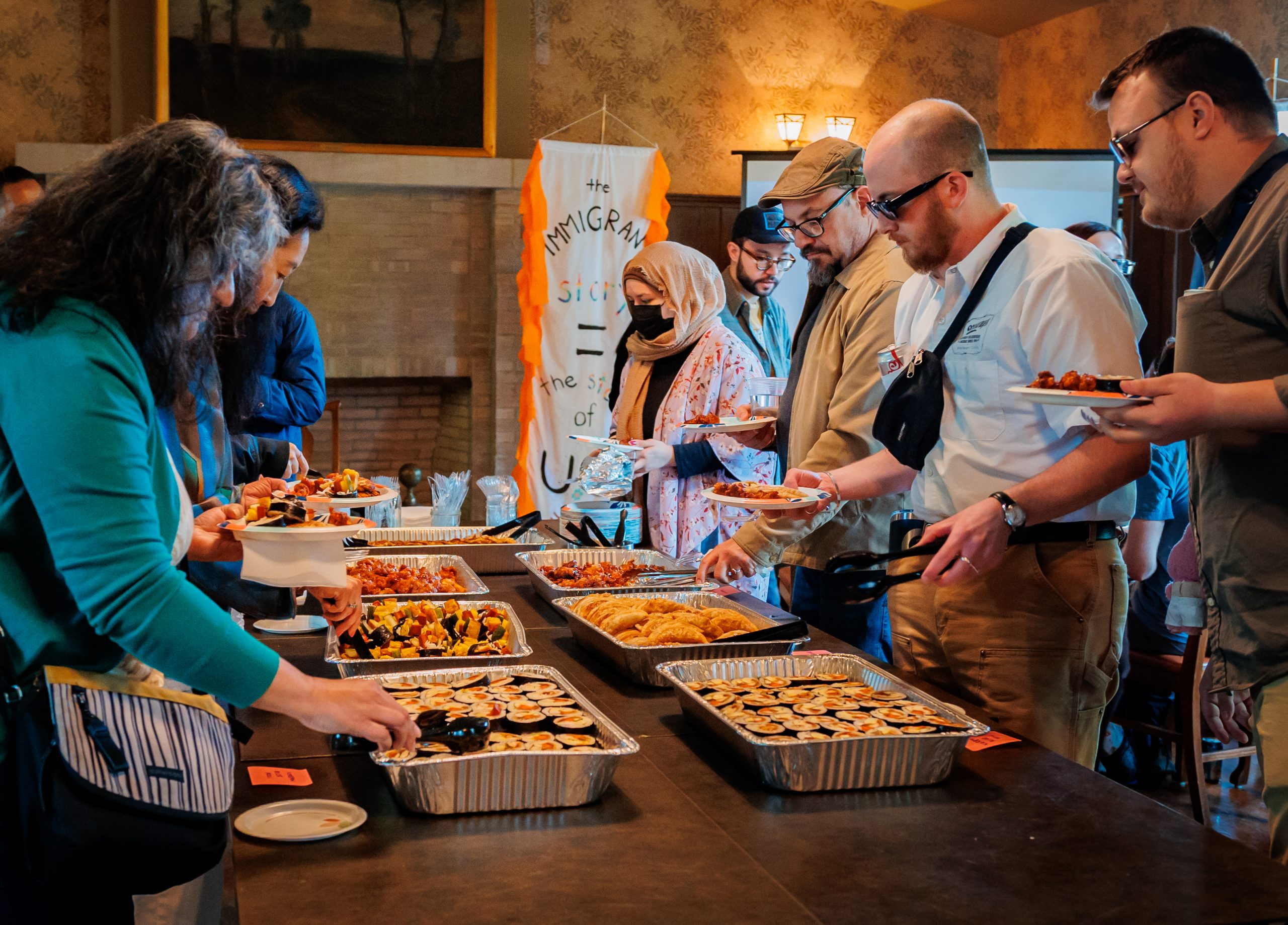 A diverse group of people serves themselves food from trays of various dishes at a buffet table during a social gathering. A banner in the background reads, Immigrants are the strength of the USA.