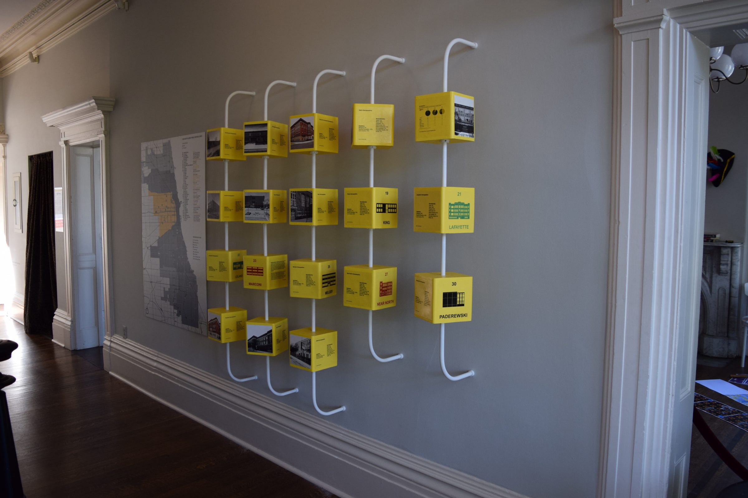 A display of yellow cubes with text and images, arranged in a grid on a gray wall with white curved supports, next to a large map in a room with wooden floors and decorative trim.