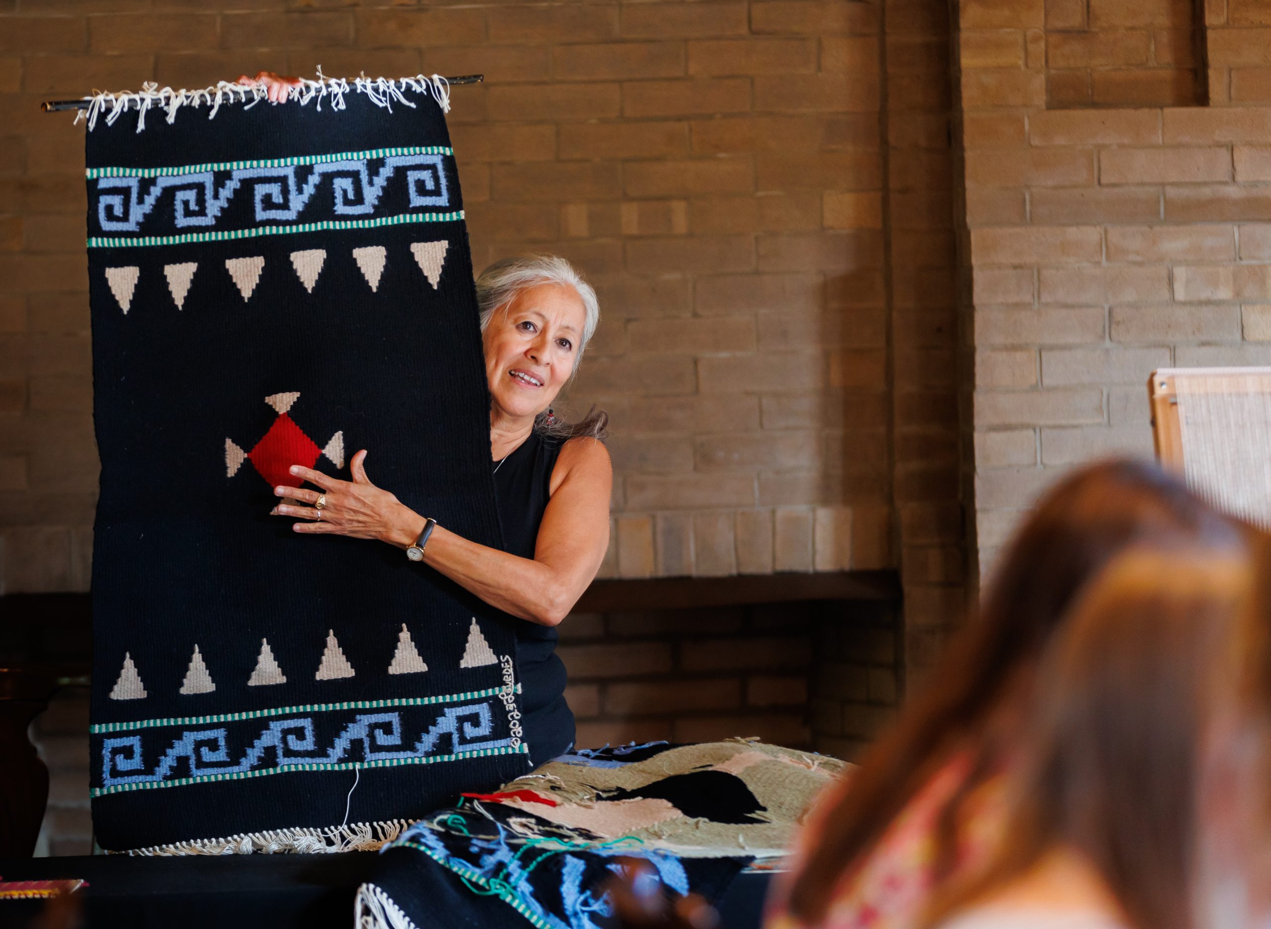 A woman with gray hair holds up a woven textile with geometric patterns in black, blue, white, and red, standing in front of a brick wall. Several people are seated in the foreground.