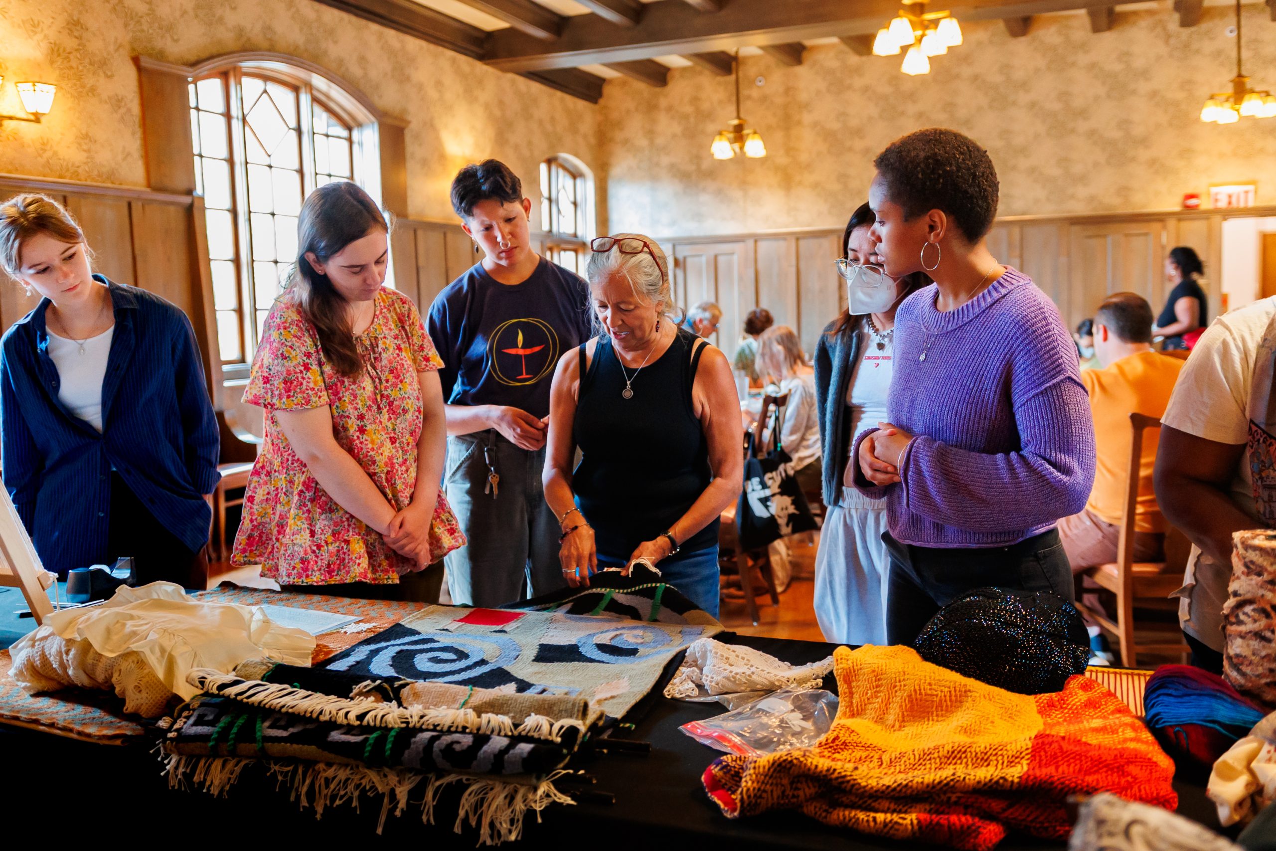 A group of people stand around a table displaying colorful textiles and crafts in a warmly lit room, attentively listening to a woman demonstrating or explaining her work. Other people are seated in the background.