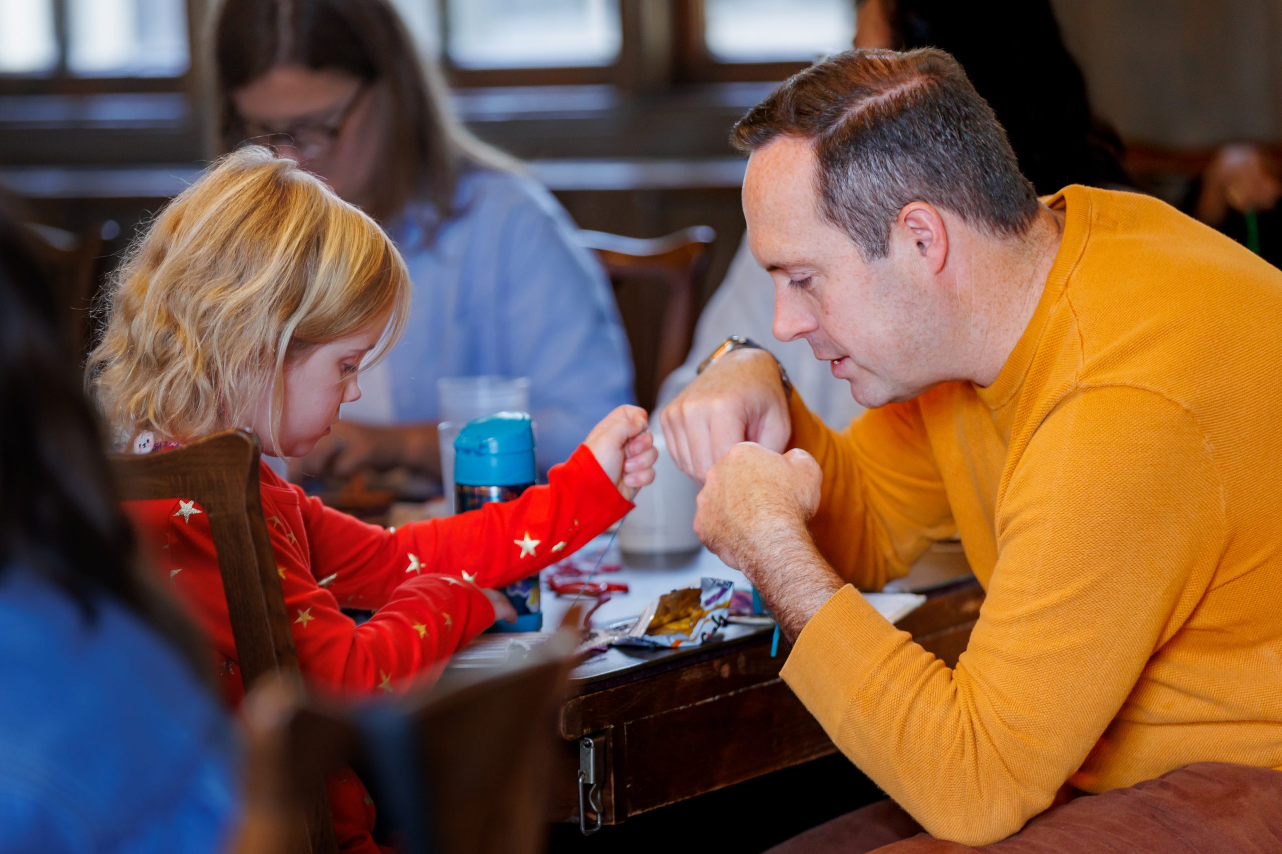A young child and an adult sit at a table, touching fists in a playful gesture. The child wears a red shirt with stars and the adult wears an orange sweater. Both are smiling and engaged with each other.