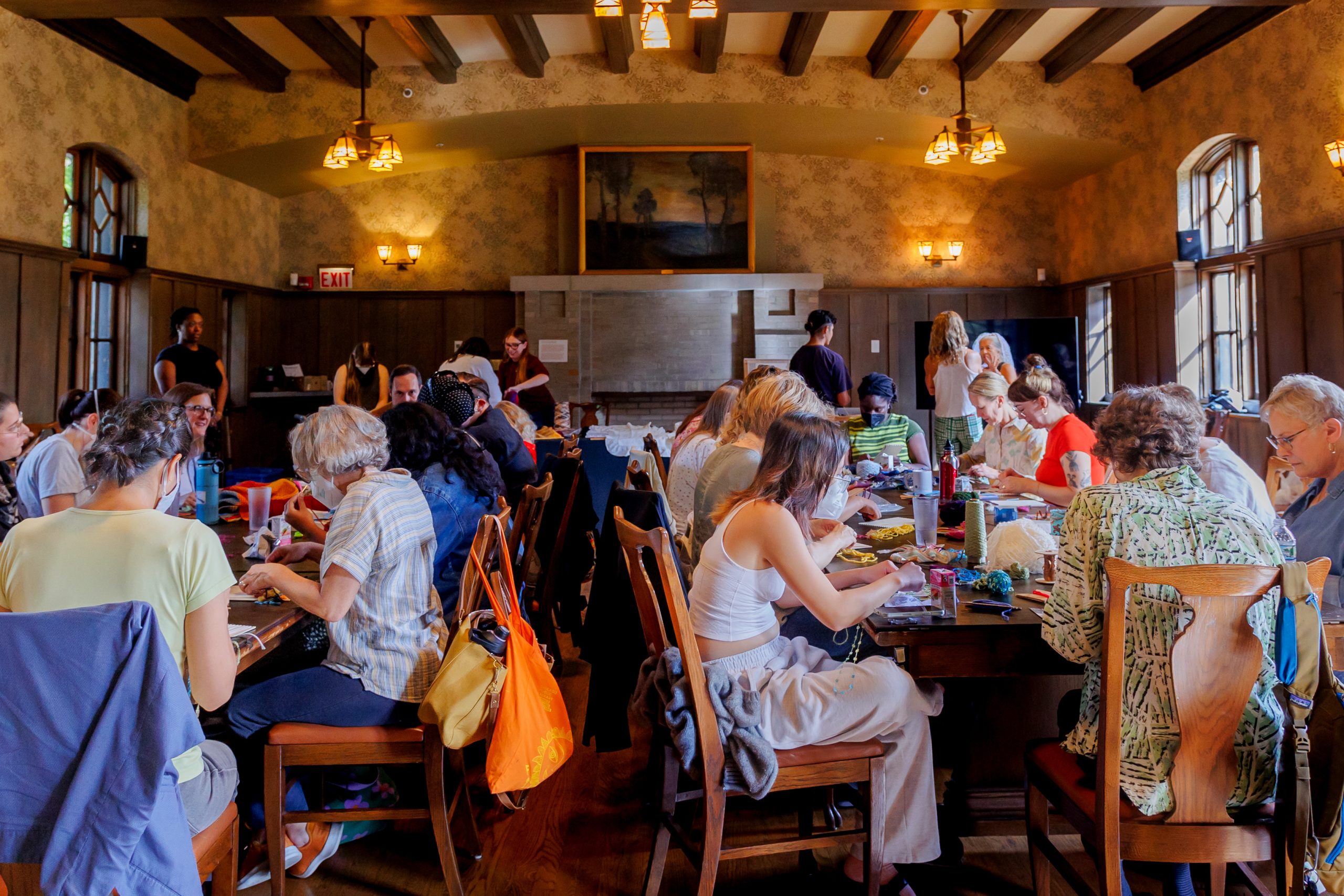 A large group of people sits around two long wooden tables in a warmly lit room, engaged in creative or collaborative activities. The room has wooden beams, hanging lights, and tall windows.