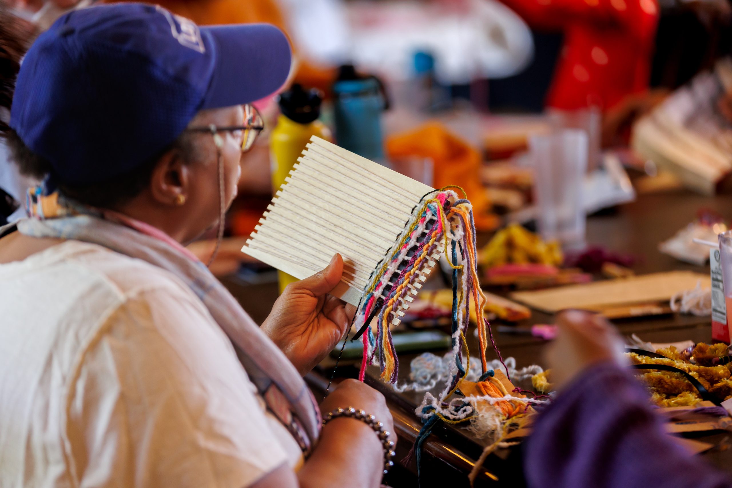 A person wearing a blue cap and glasses holds a small loom with colorful threads, participating in a group weaving or crafting activity at a table with various materials.