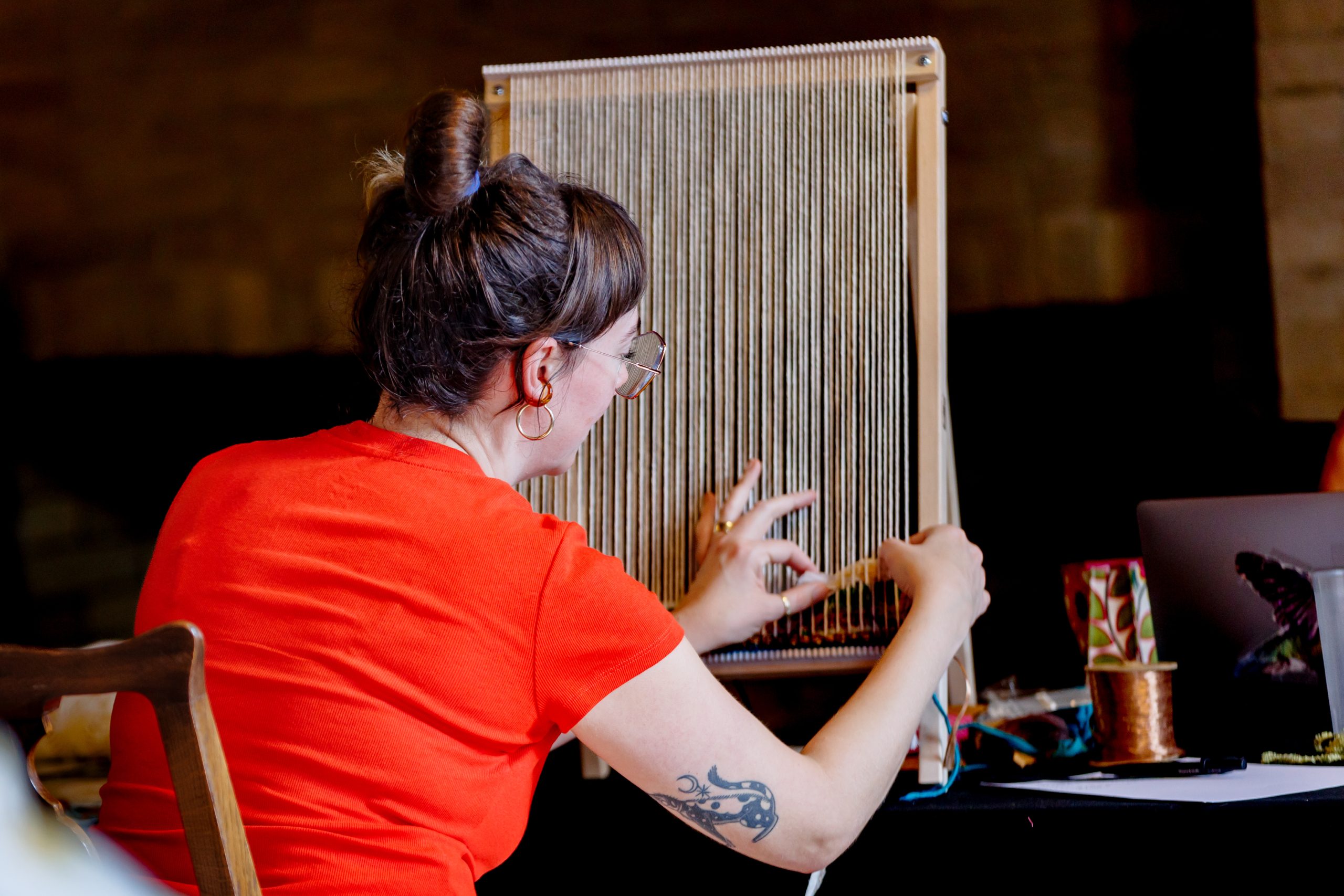 A person with a bun hairstyle and tattoo on their arm weaves on a vertical loom, wearing a bright orange shirt, seated at a table with crafting materials nearby.