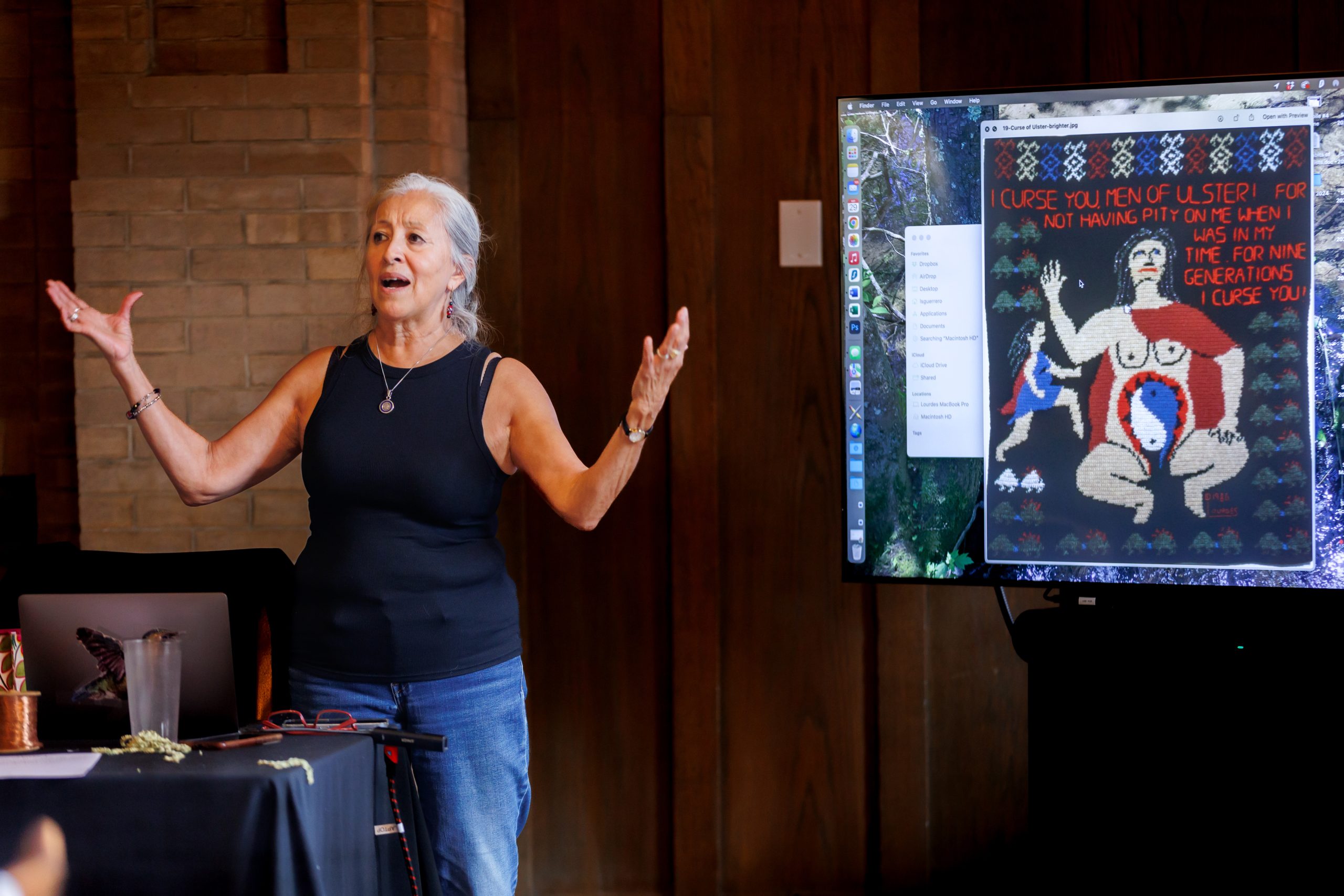 A woman with gray hair stands with arms open, speaking beside a large screen displaying colorful artwork that includes text and figures. The background features wooden walls and a brick fireplace.