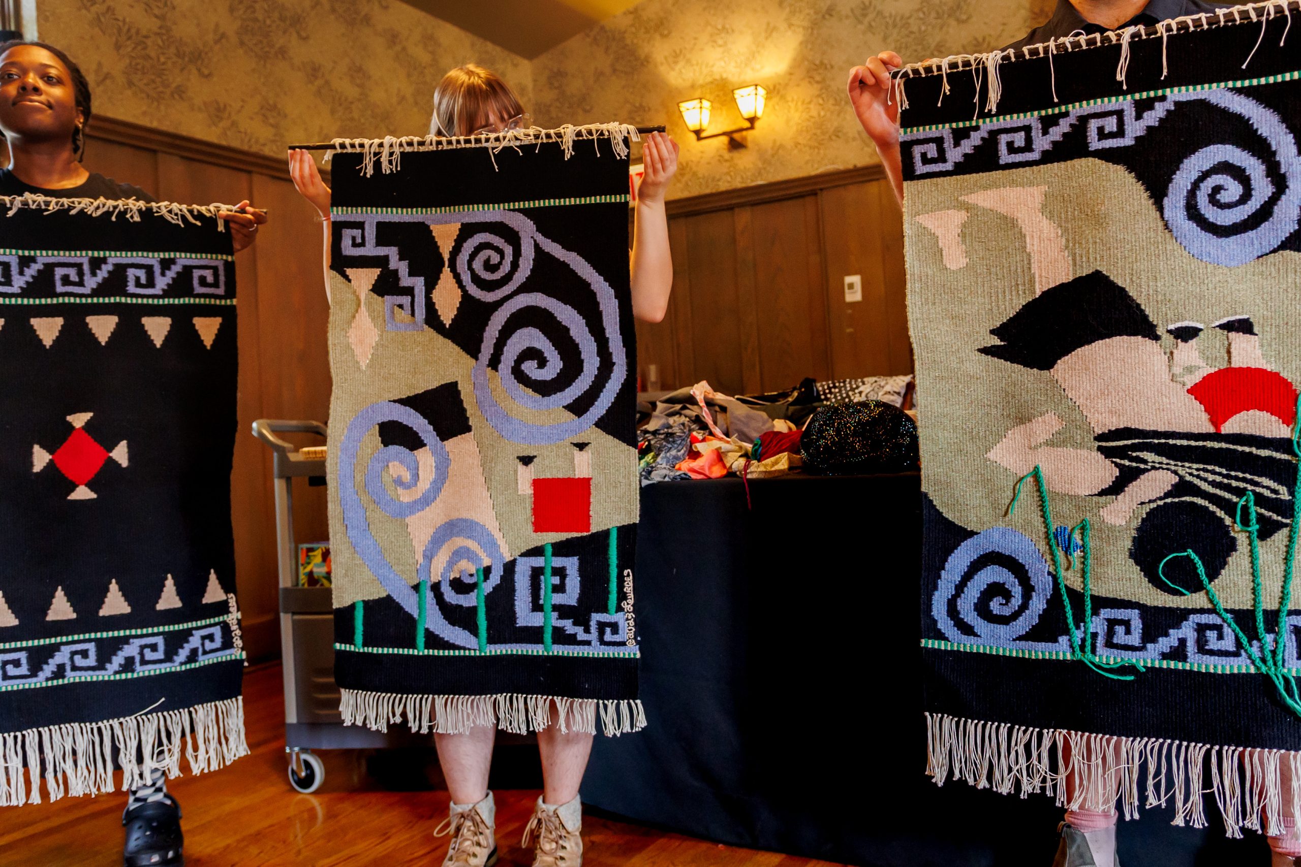 Three people hold up colorful, abstract woven tapestries with geometric and spiral patterns. Only their arms and legs are visible; the focus is on the intricate designs of the textiles.