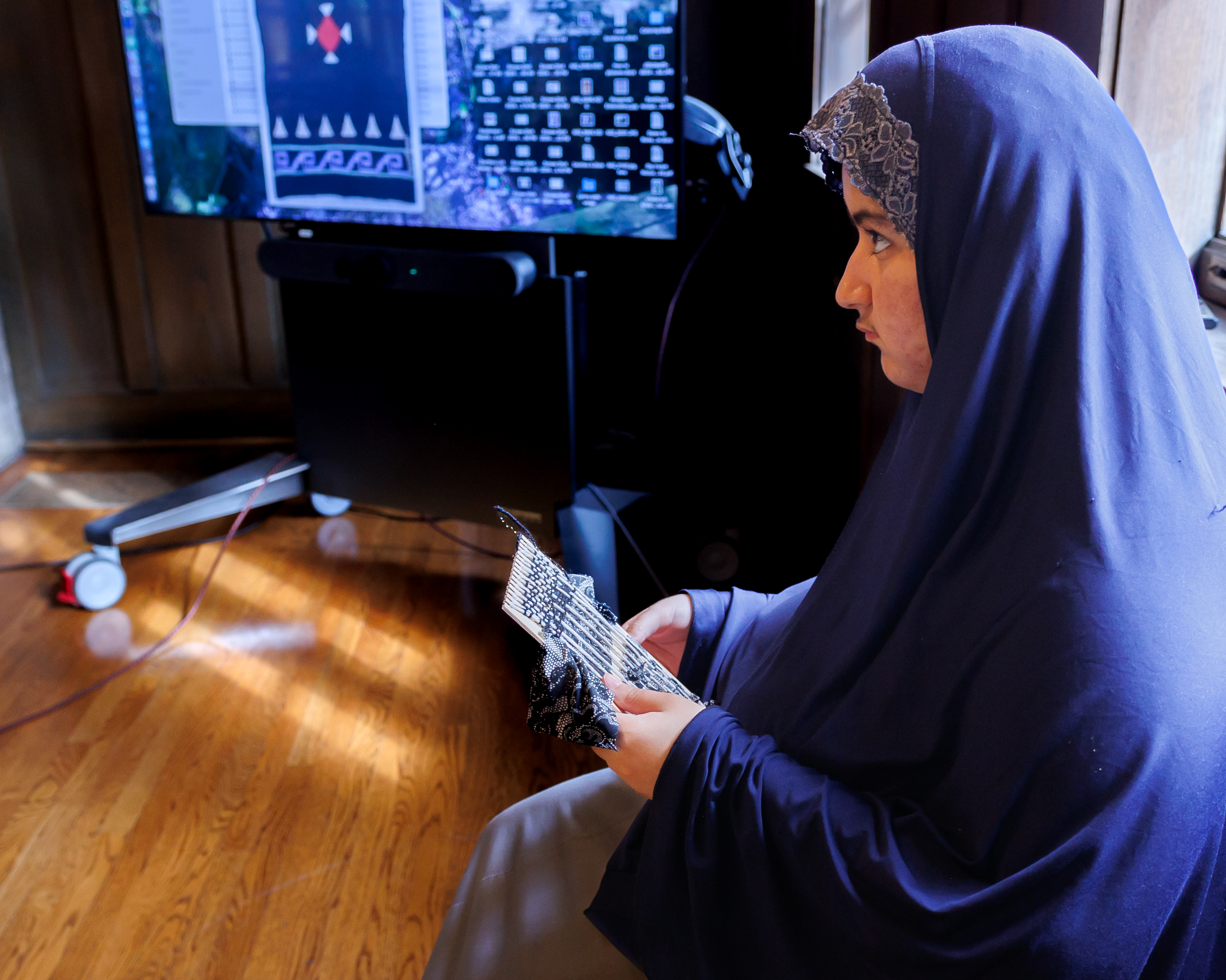 A woman in a navy hijab sits on a wooden floor, holding prayer beads and a paper. Behind her, a large monitor displays computer icons and graphics.