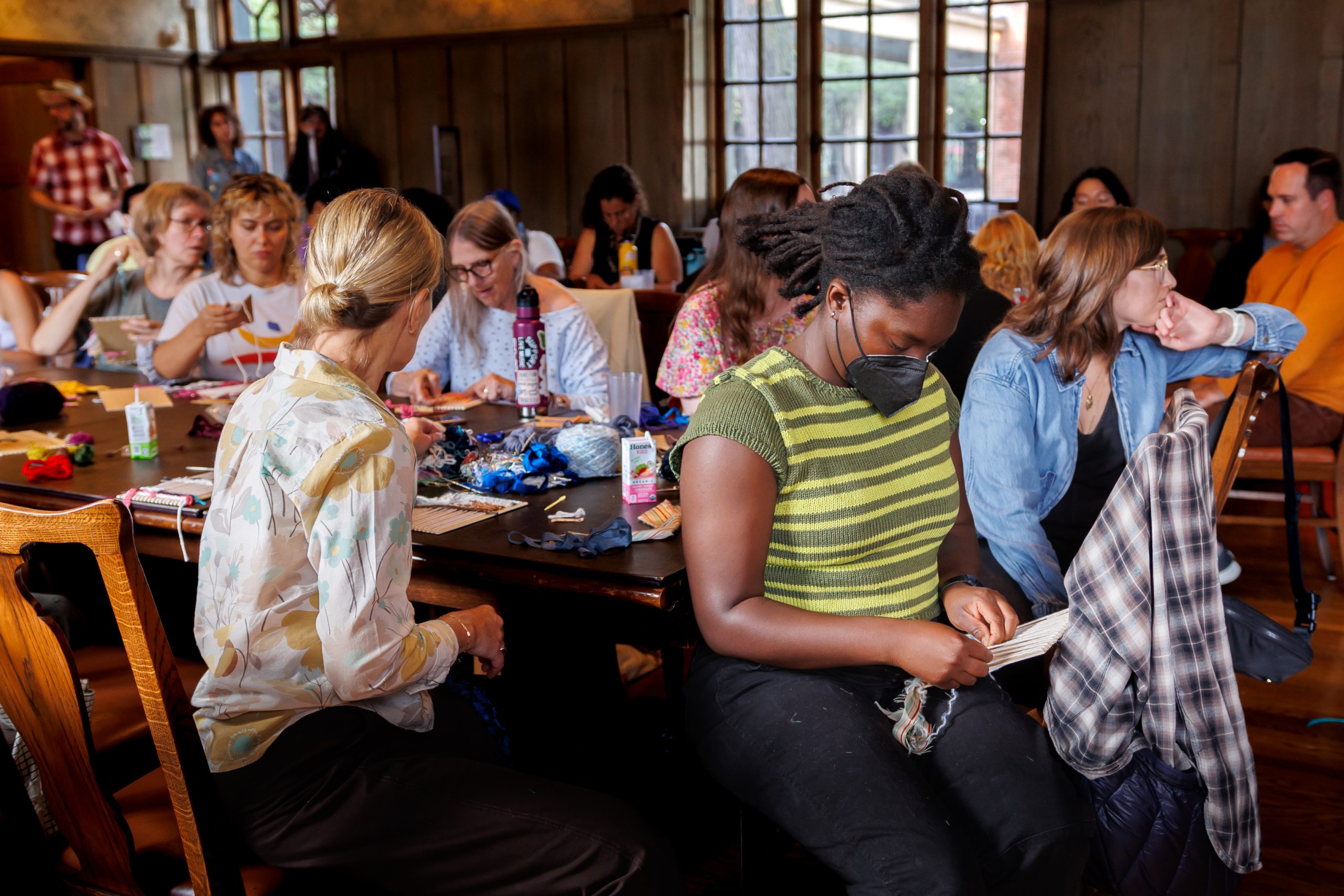 A group of people sit at tables in a well-lit room, focused on crafts or sewing projects. Art supplies and fabric are scattered on the tables, and one person in the foreground wears a face mask while working.
