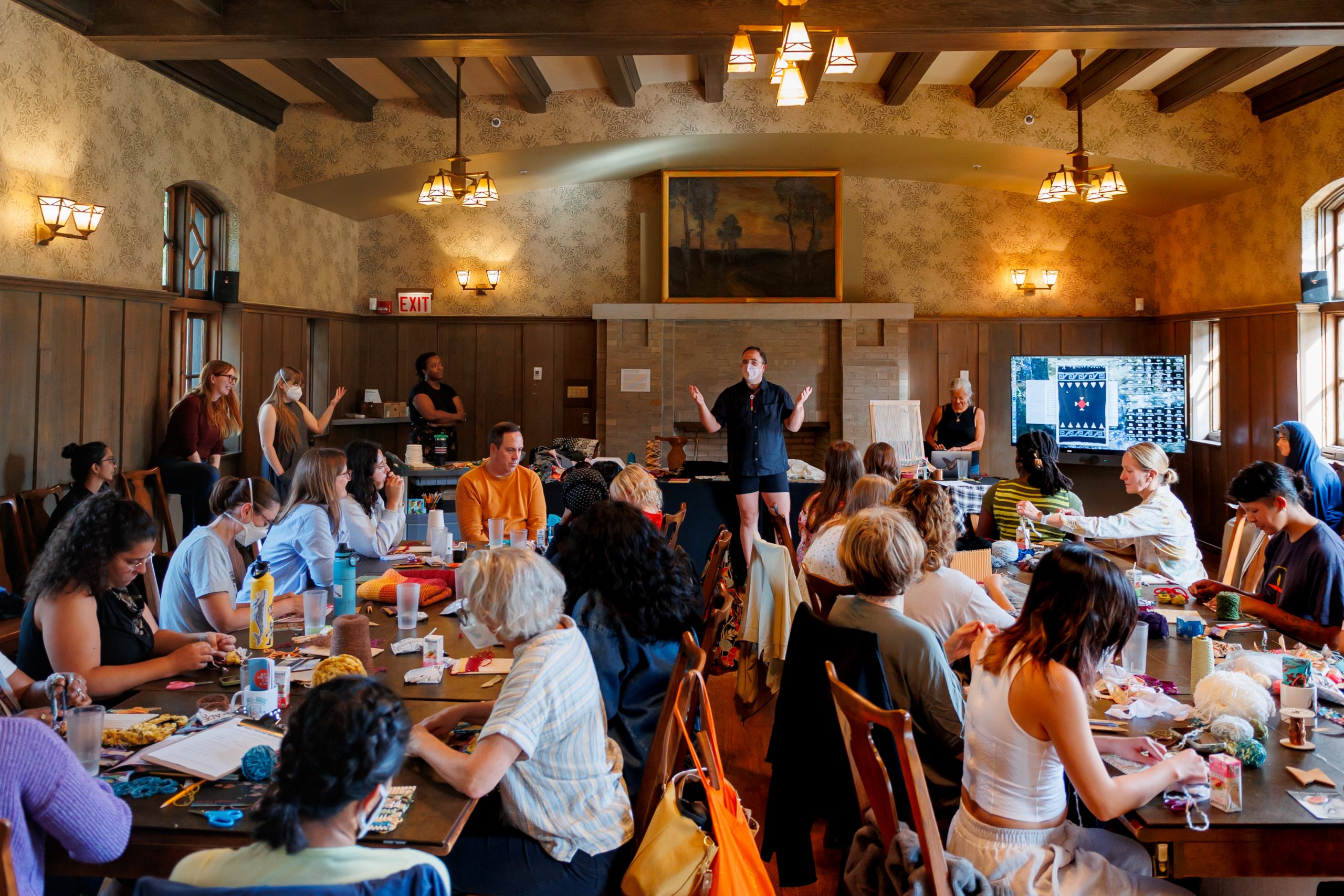 A group of people sit around a large table in a wood-paneled room, listening to a person speaking at the front near a presentation screen. Some attendees are taking notes or working on crafts.