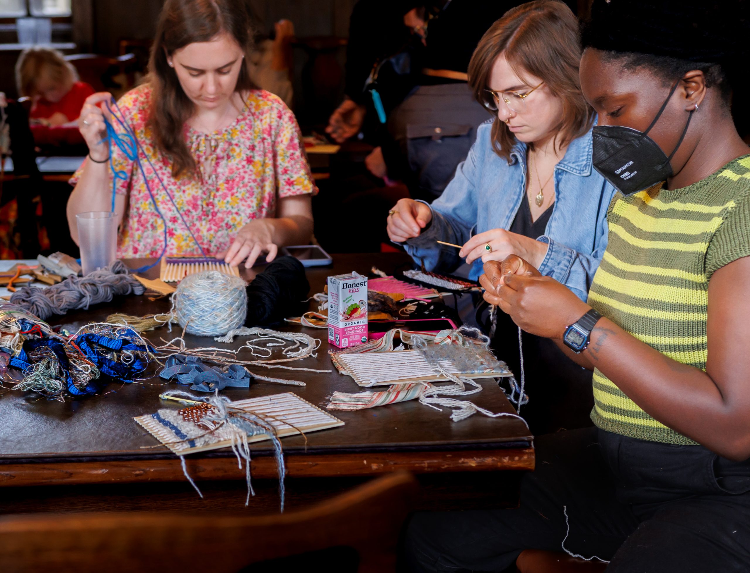 Three women sit at a table covered with yarn, weaving supplies, and drinks, focused on crafting projects. One wears a mask, and all are busy with their hands, creating textile art in a communal, indoor setting.