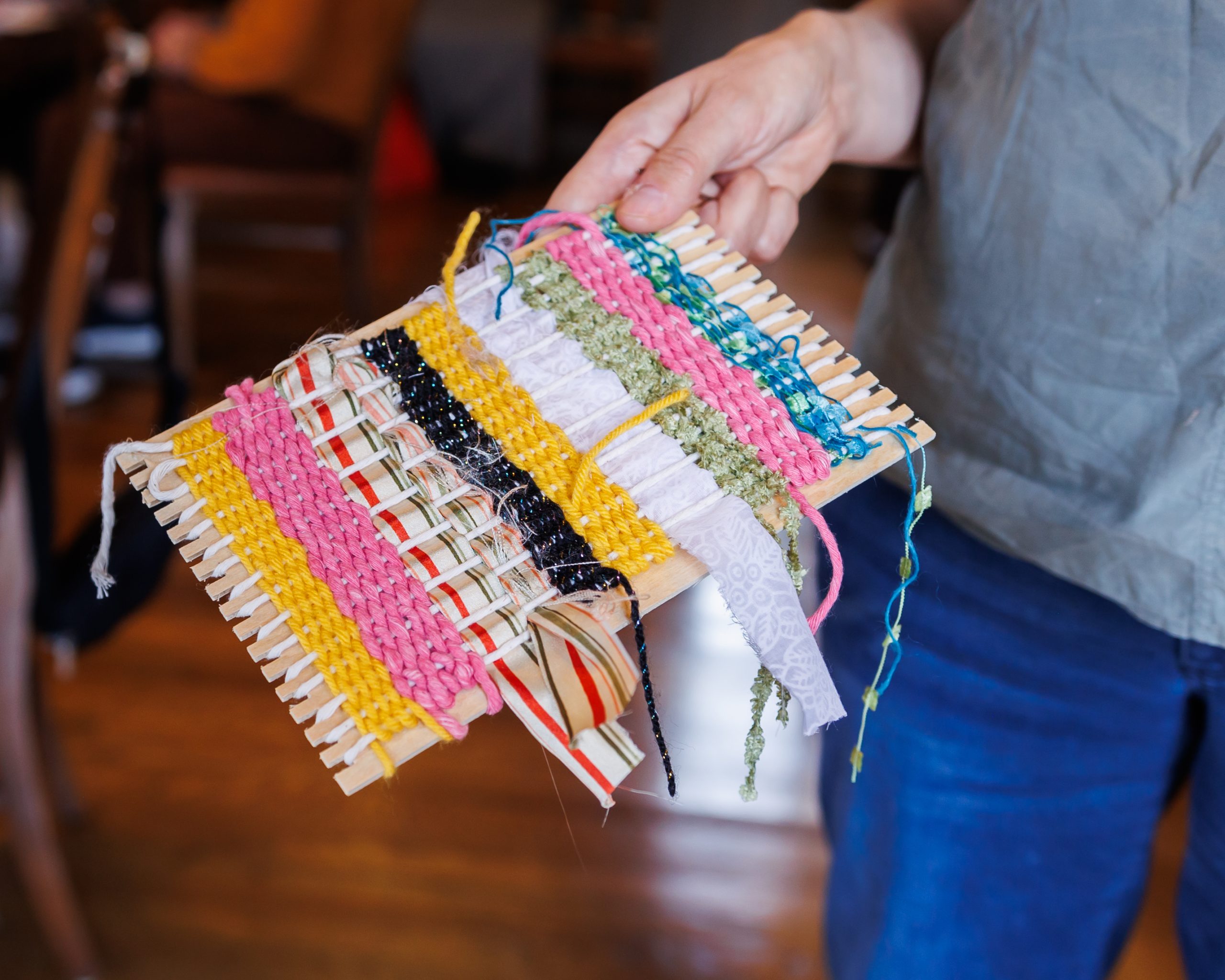 A person holds a small wooden loom with colorful woven threads and ribbons in yellow, pink, white, blue, green, and black. The loom is partially completed and the person’s hand is visible.