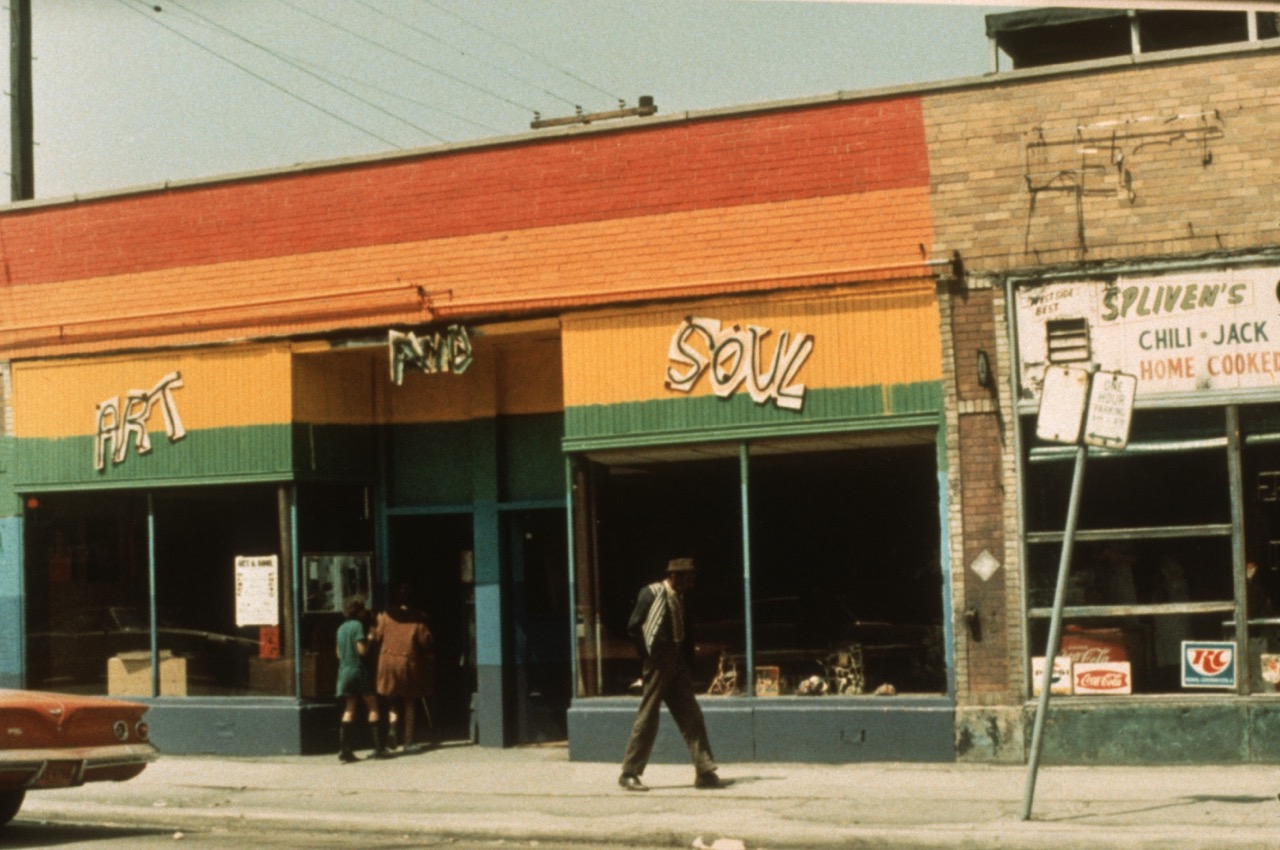 A colorful storefront labeled “Art and Soul” with green, orange, and yellow stripes. Two people enter the shop, while a man walks by. Next door is a diner sign reading “Spliven’s Chili, Jack, Home Cooked.”.