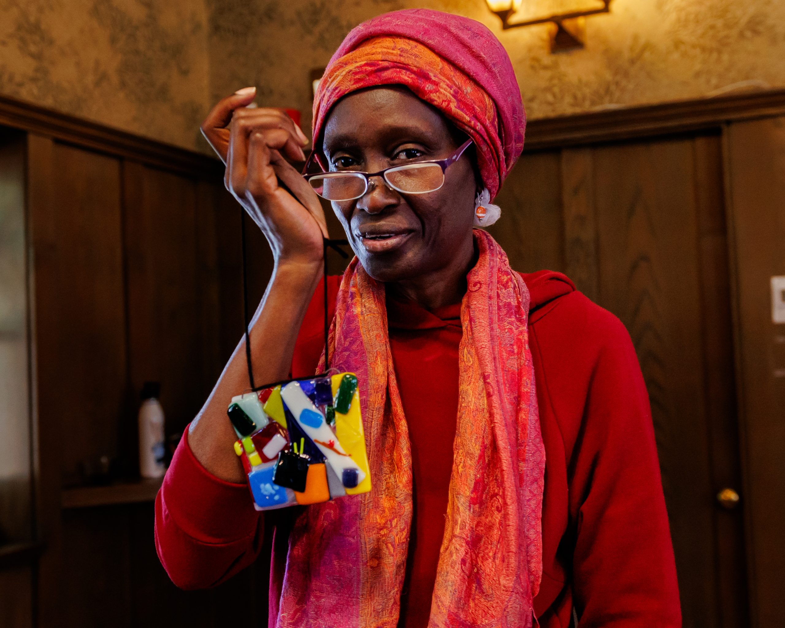 A woman in a red outfit and headscarf smiles while holding up a colorful, abstract piece of art and looking over her glasses inside a warmly lit room with wooden walls.