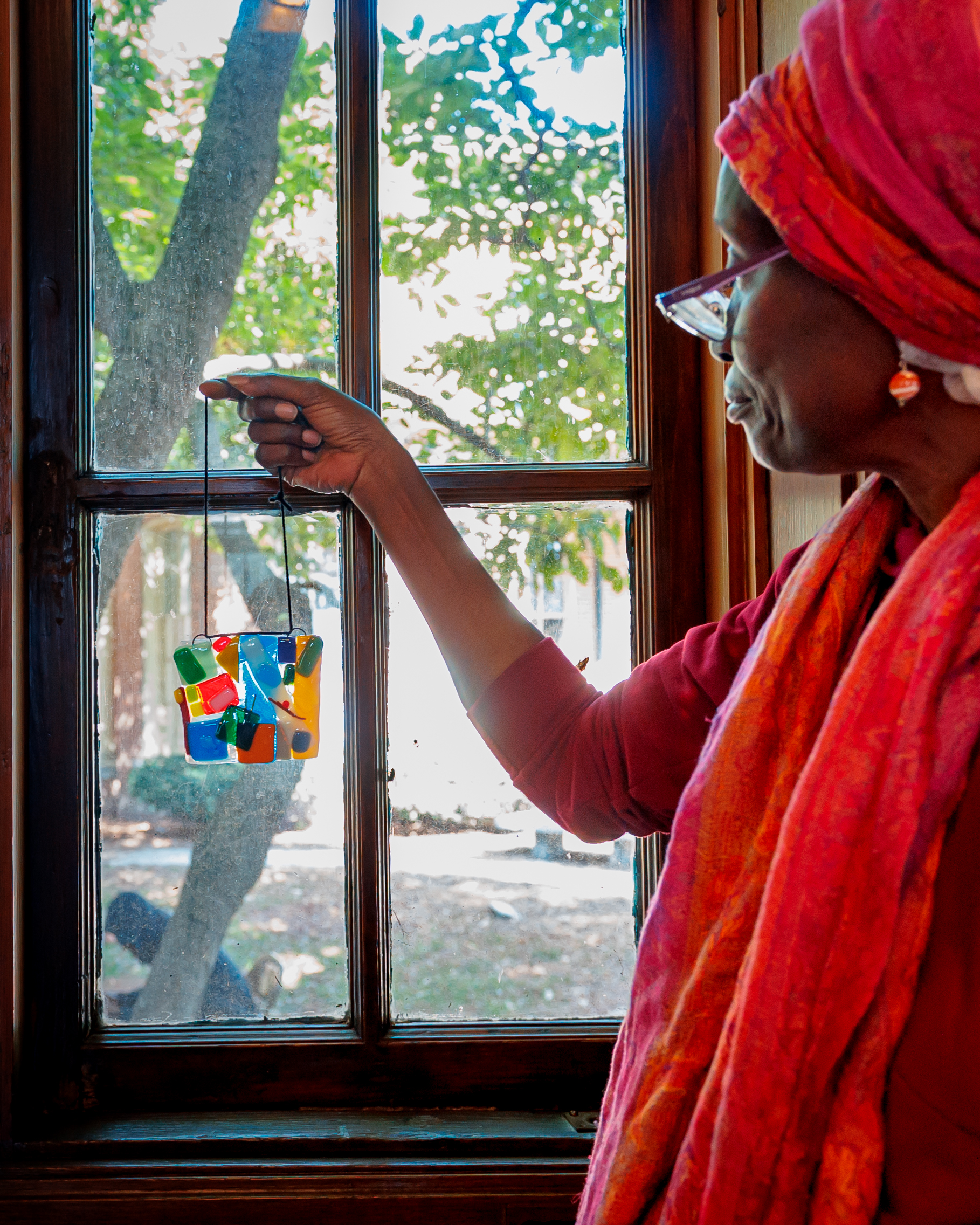 A woman in a pink headscarf and glasses holds up a colorful piece of stained glass near a wooden window, with trees and sunlight visible outside.