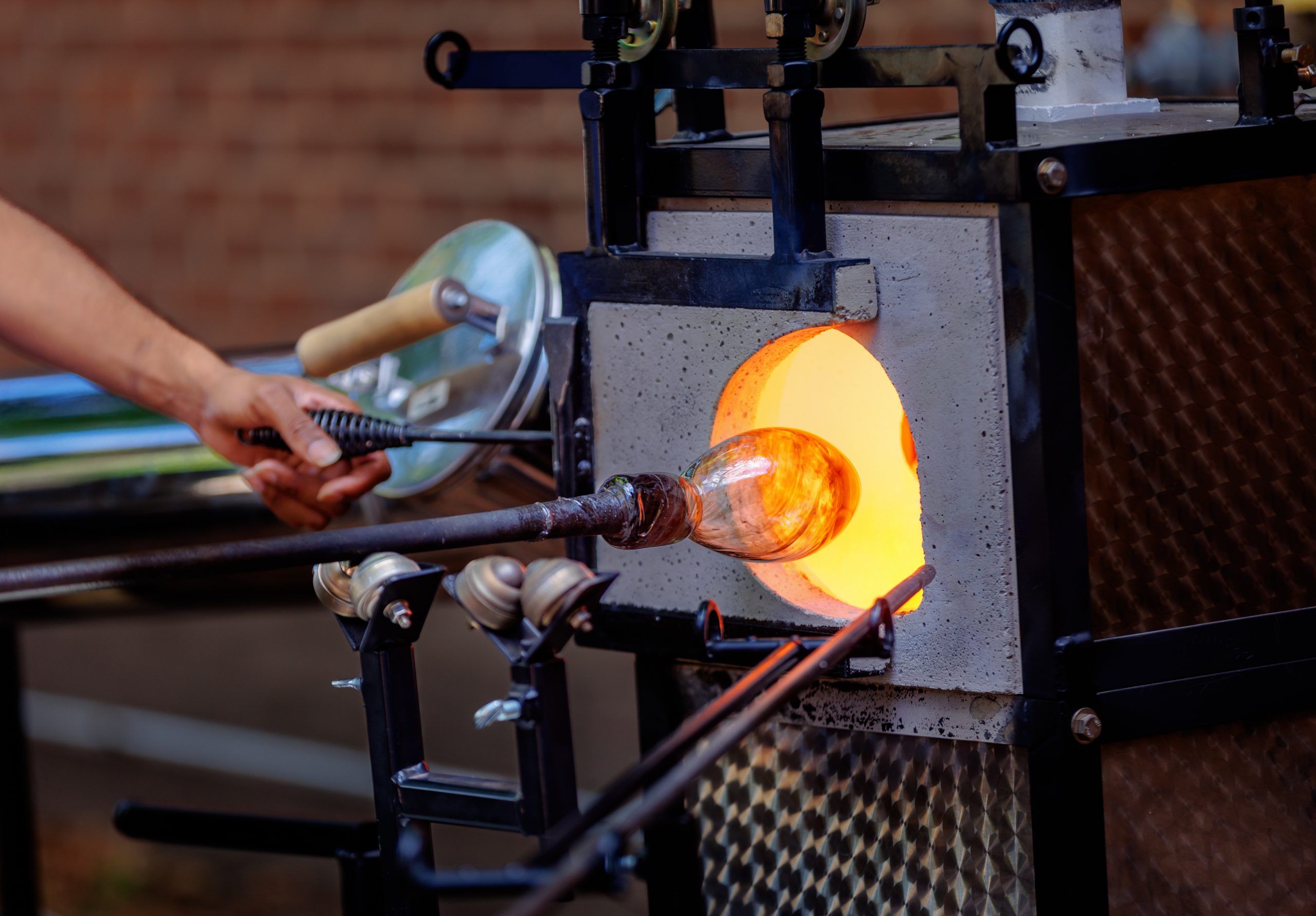 A person shapes molten glass with a tool at the opening of a hot furnace, creating a glass object on the end of a metal rod. The workspace and equipment are visible in the background.