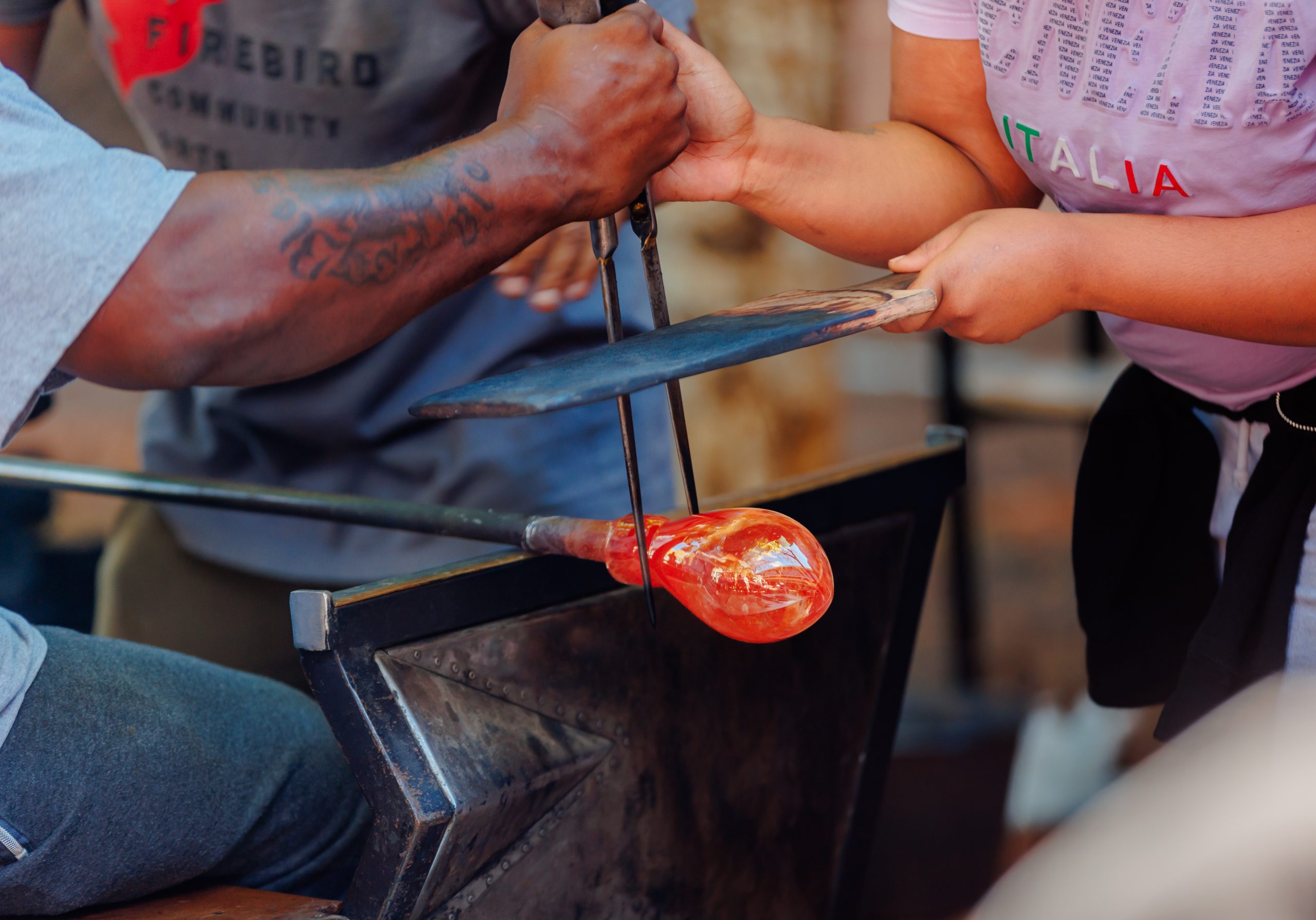 Two people work together to shape molten glass using tools; one holds the glass while the other uses metal tongs and a paddle to form it. The orange-hot glass is being crafted on a workbench.