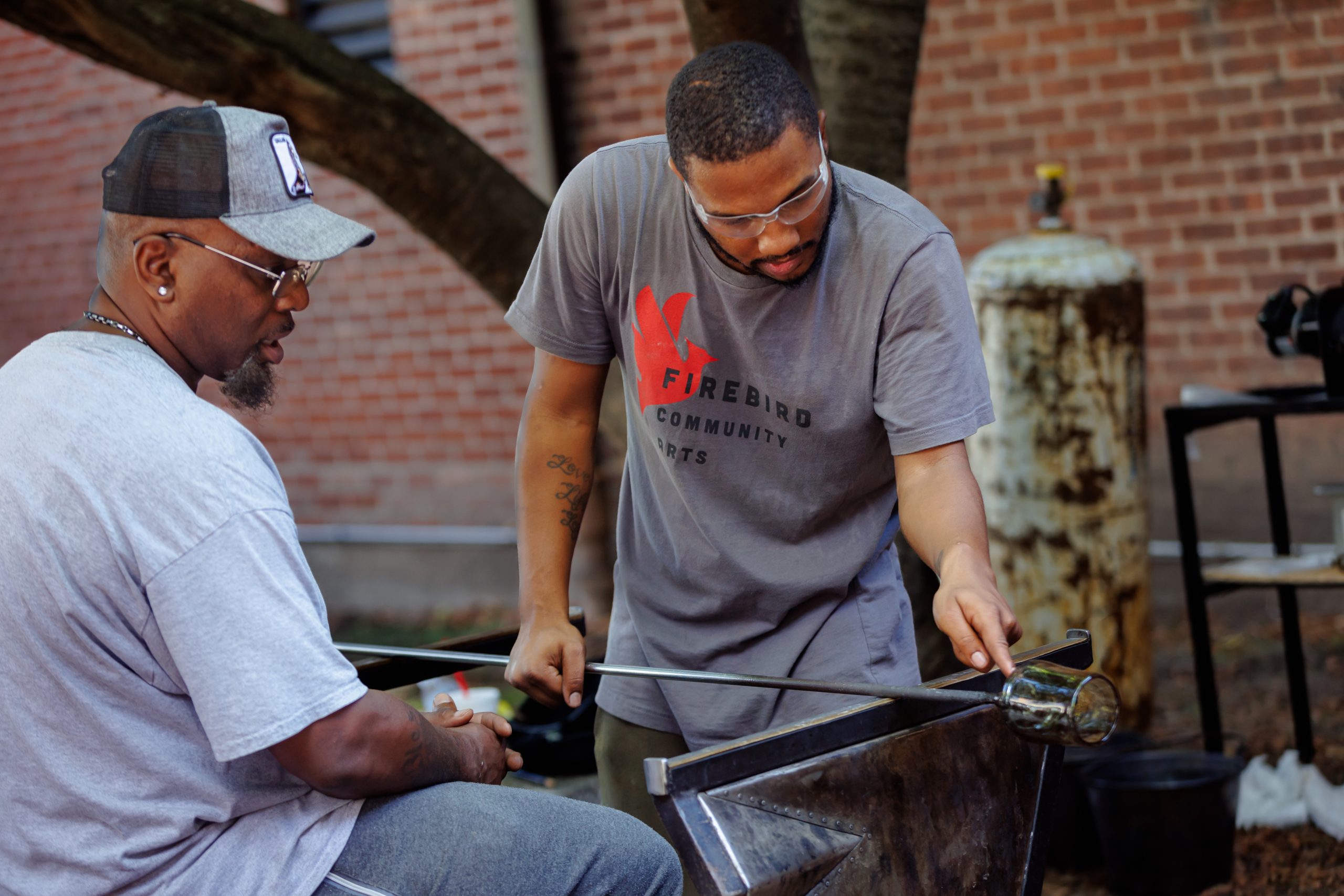 Two men work together on a glassblowing project outdoors. One holds a rod with molten glass while the other shapes it. They are focused, wearing casual clothes, with a brick wall and trees in the background.