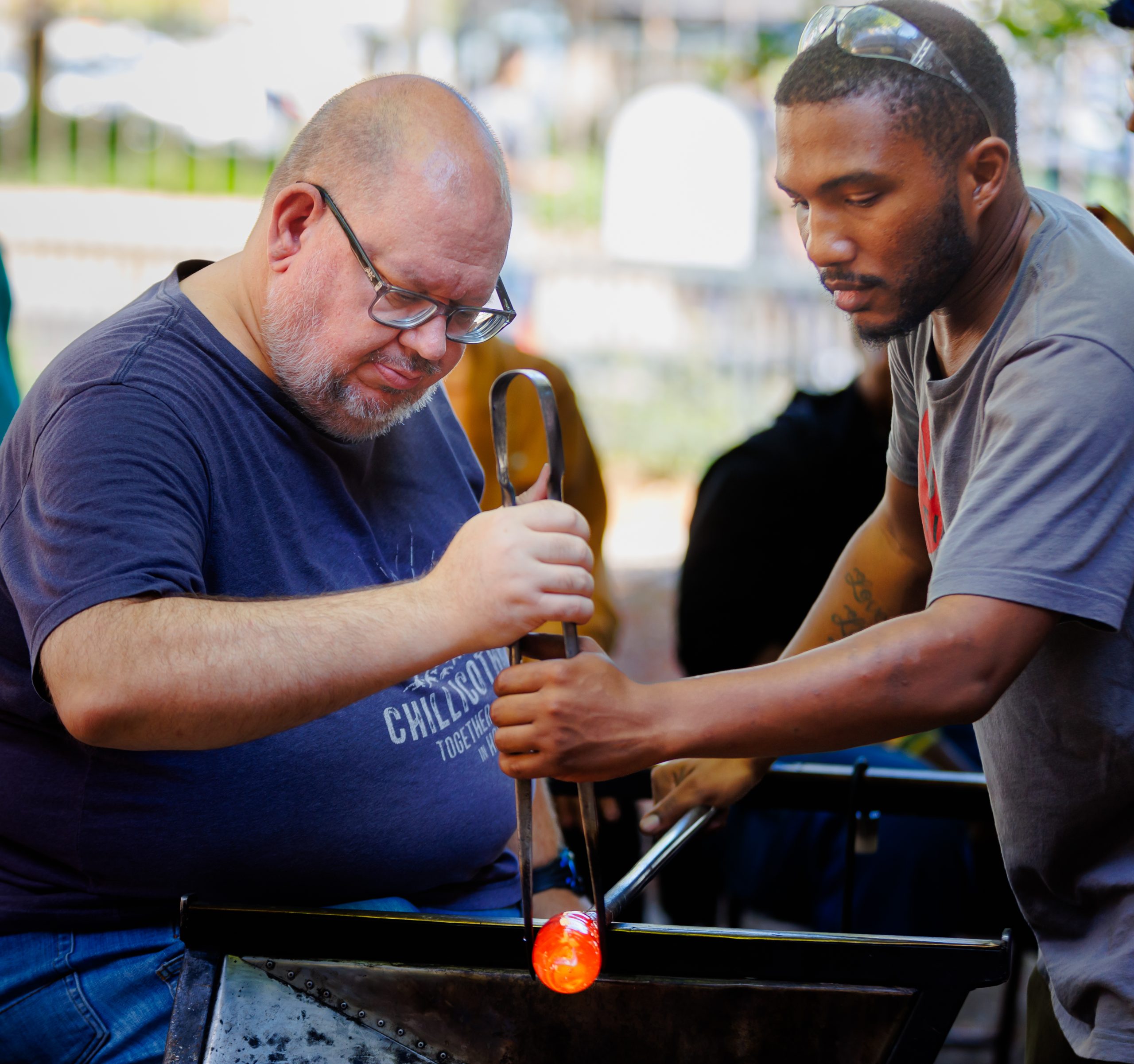 Two men work together on glassblowing; one holds tongs gripping molten glass while the other assists, both focused on shaping the glowing orange material. They are outdoors with blurred nature in the background.