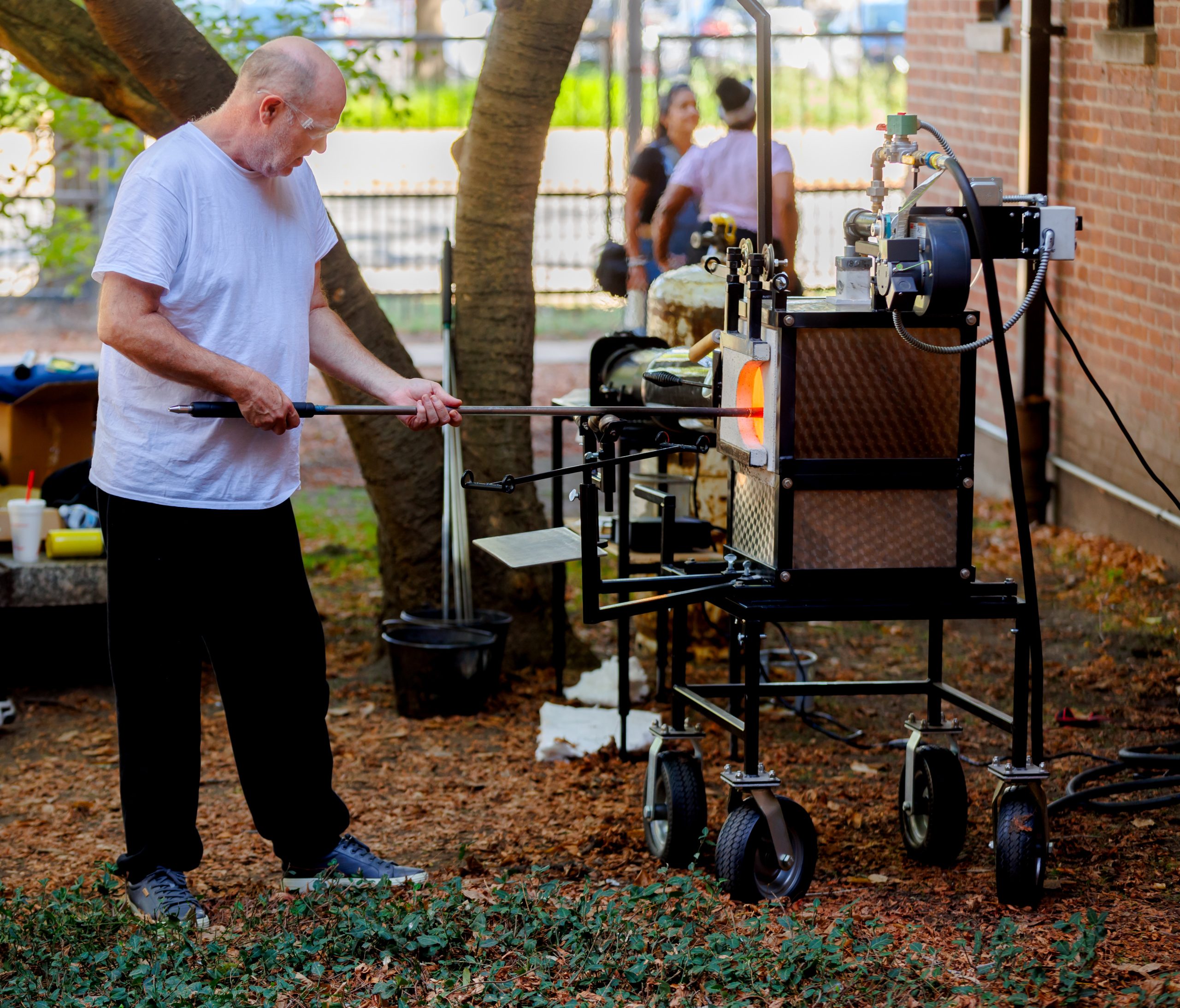 A man in a white shirt and black pants shapes glass using a long rod at an outdoor glassblowing furnace. Trees and a brick building are in the background, with people visible further away.