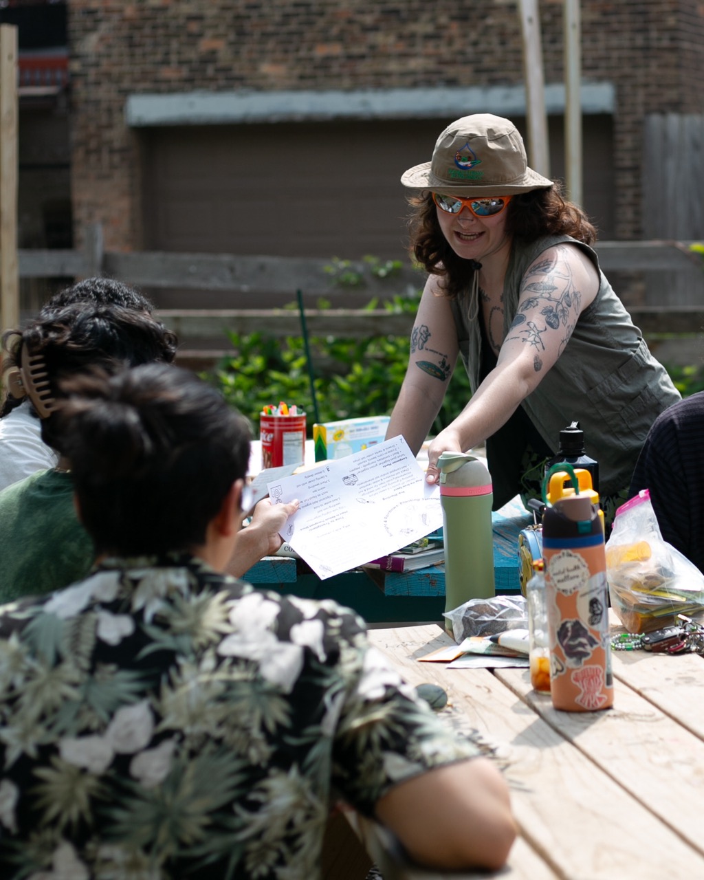 A person wearing a hat and sunglasses hands a paper to a group of people sitting at an outdoor wooden table covered with water bottles and art supplies, with a brick building in the background.