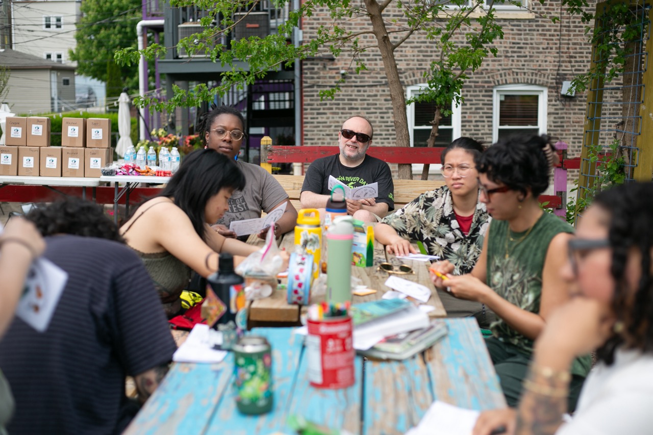 A diverse group of people sit around a colorful outdoor table, engaged in conversation and activities, with food, drinks, and art supplies on the table. Boxes and beverages are stacked in the background.