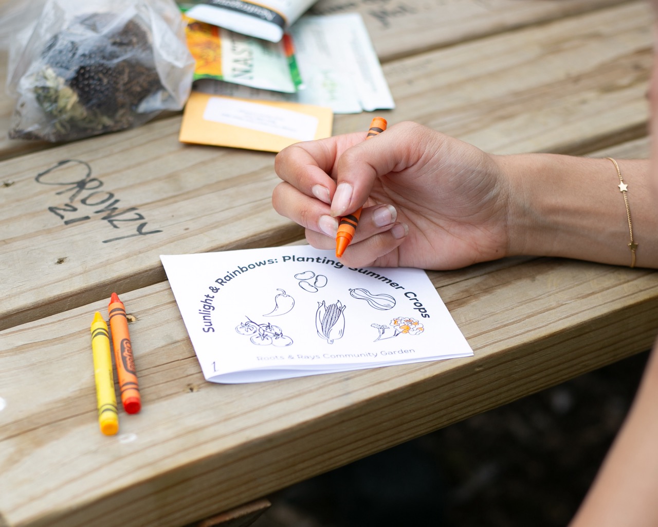 A person colors a gardening activity sheet titled Sunlight, Rainbows: Planting Summer Crops with crayons at a wooden table, with seed packets and crayons nearby.