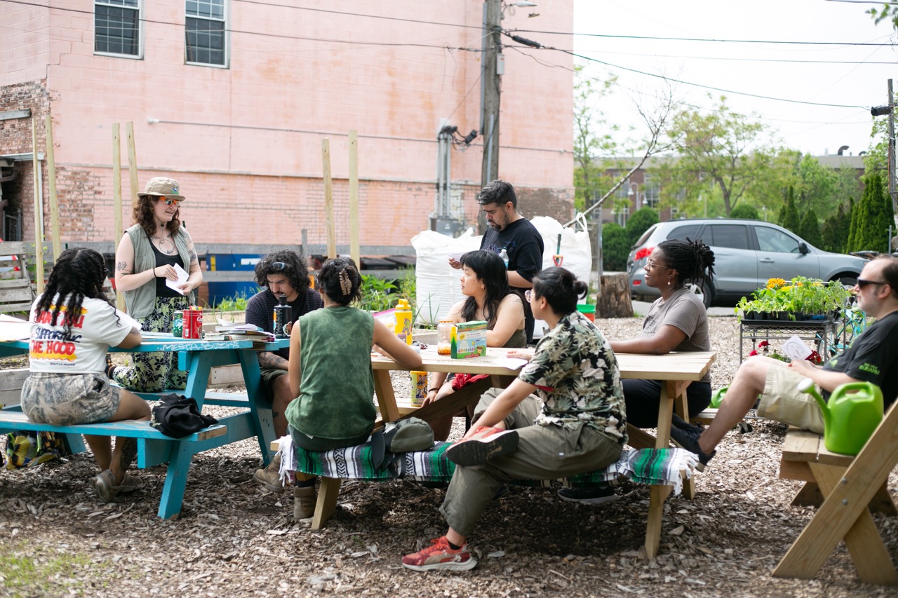 A group of people sit around picnic tables outdoors, listening to a woman standing and speaking. Gardening supplies and plants are nearby, and a car is parked in the background. It appears to be a community garden gathering.