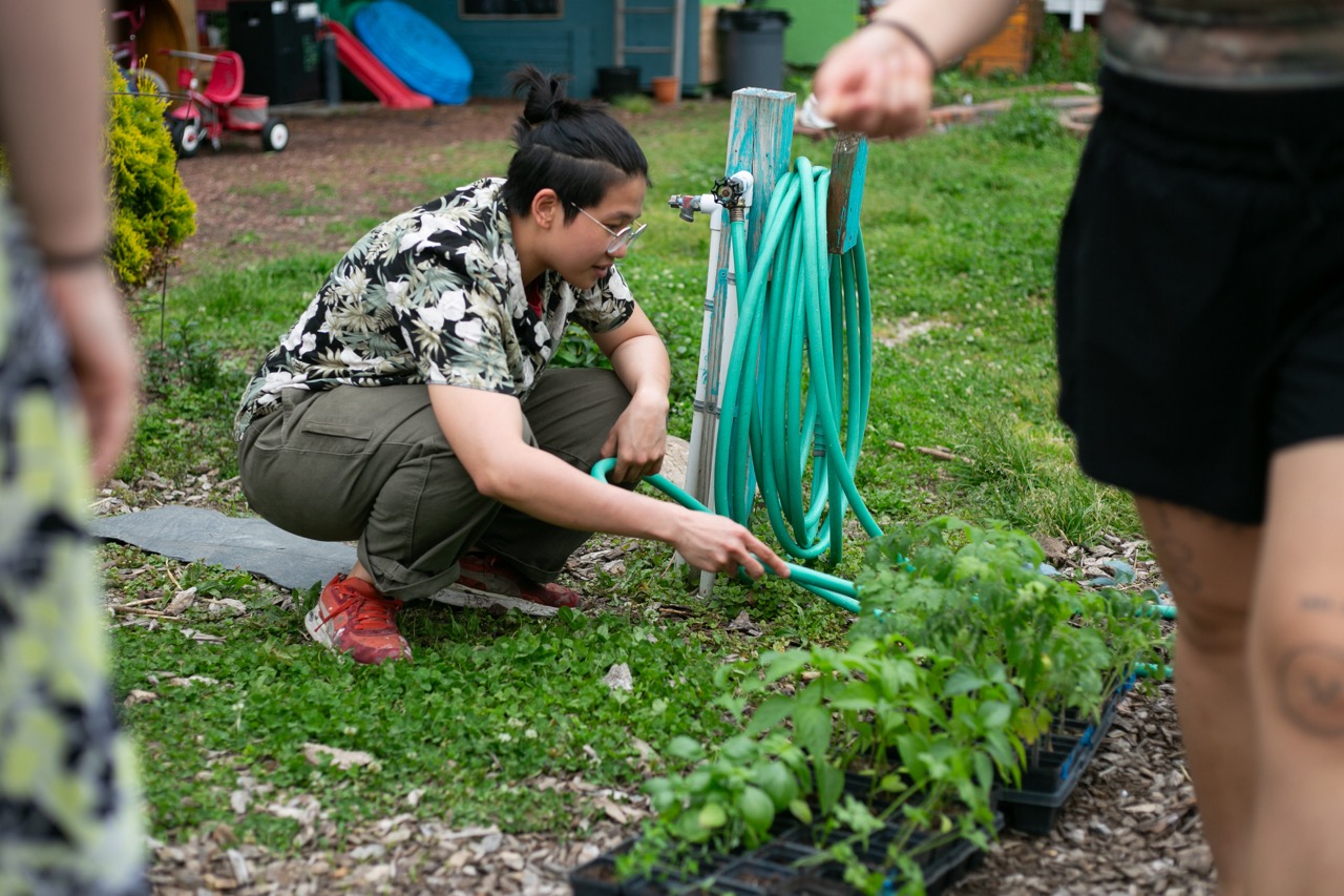 A person crouches beside a garden hose, watering seedlings in trays outdoors. They wear glasses, a floral shirt, and red shoes. Another person, partially out of frame, stands nearby.