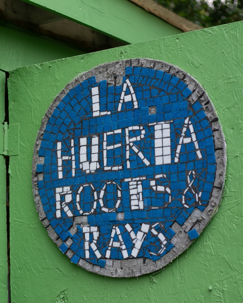 A circular mosaic sign on a green wall reads La Huerta Roots & Rays in white and blue tiles.