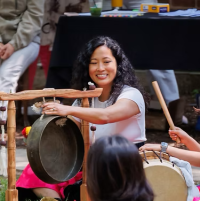 A woman smiles while striking a gong with a mallet during an outdoor drumming activity, surrounded by children playing drums.