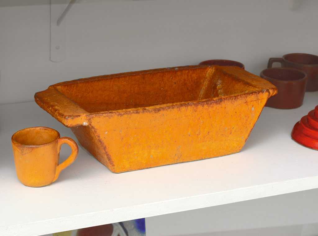 A rectangular, orange ceramic baking dish with a rough texture sits on a white shelf next to a matching orange mug. Other red and orange ceramic cups and objects are partially visible in the background.