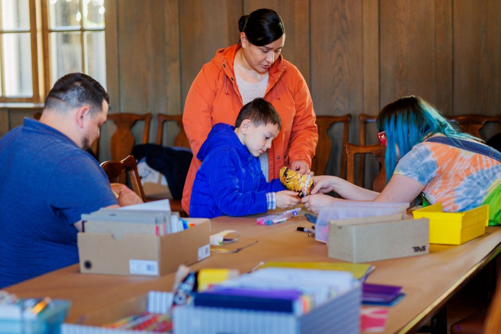 Four people sit and stand around a table with craft supplies. An adult helps a child in a blue jacket with a craft, while another adult and child work together. Boxes of supplies are scattered on the table in a cozy, wood-paneled room.