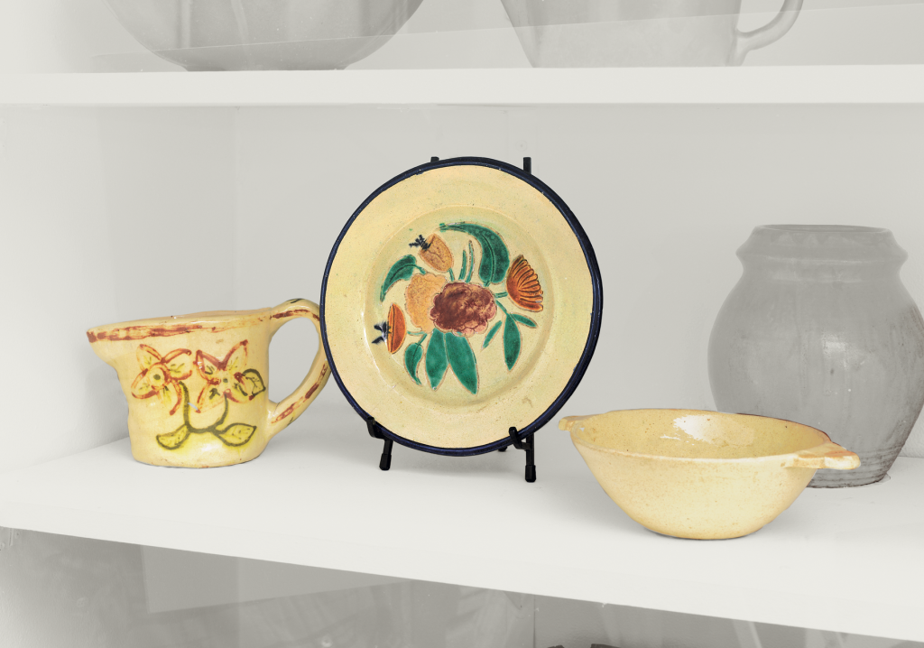 A display shelf with three pieces of pottery: a cup with yellow and red floral designs, a plate with painted flowers on a stand, and a shallow yellow bowl. A plain clay pot is in the background.