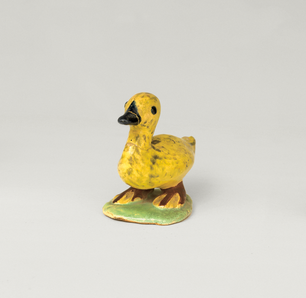 A small yellow duck figurine with a black beak, standing on a green base against a plain light gray background.