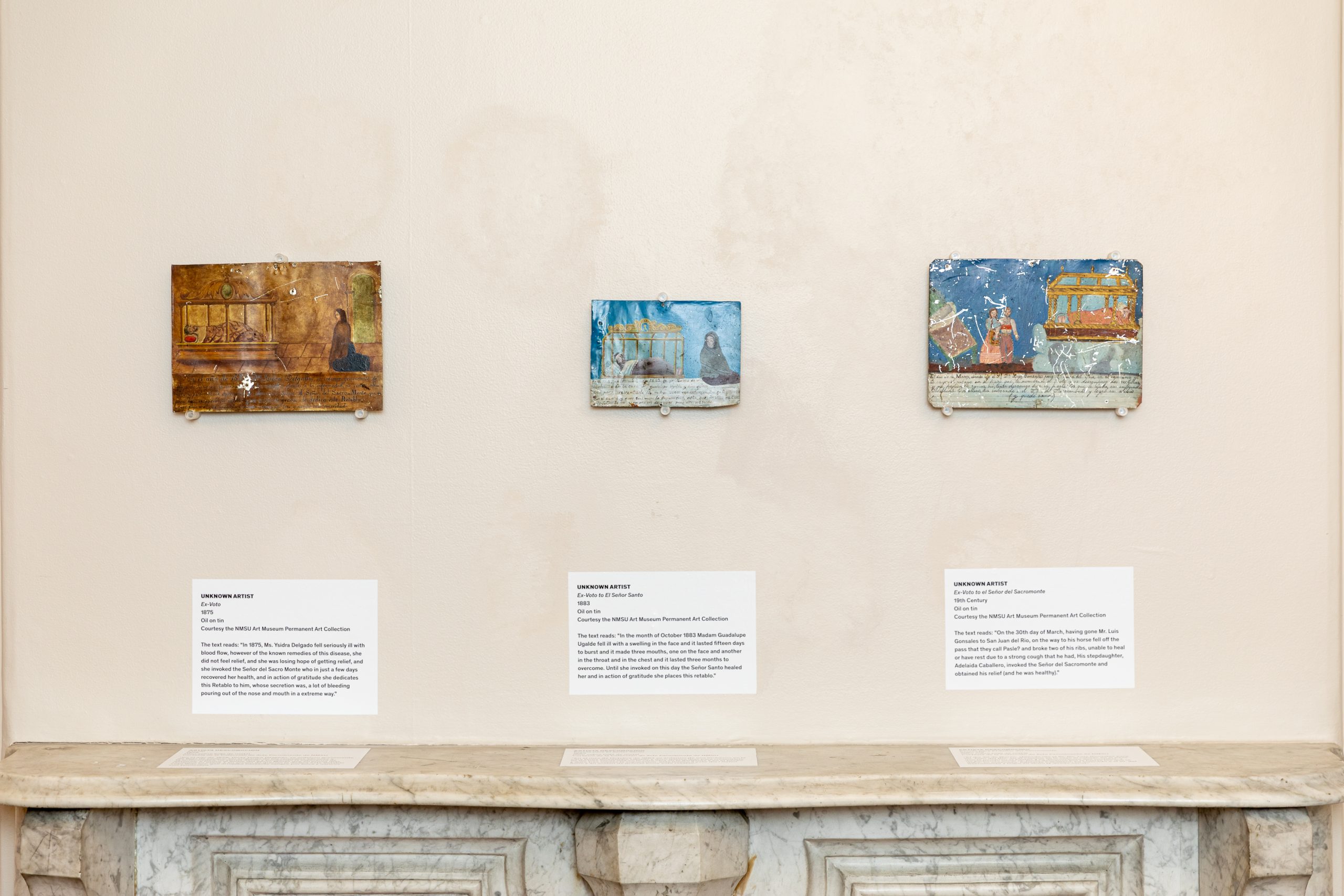 Three small rectangular paintings with informative labels are displayed on a plain wall above a white marble mantelpiece in a gallery setting.