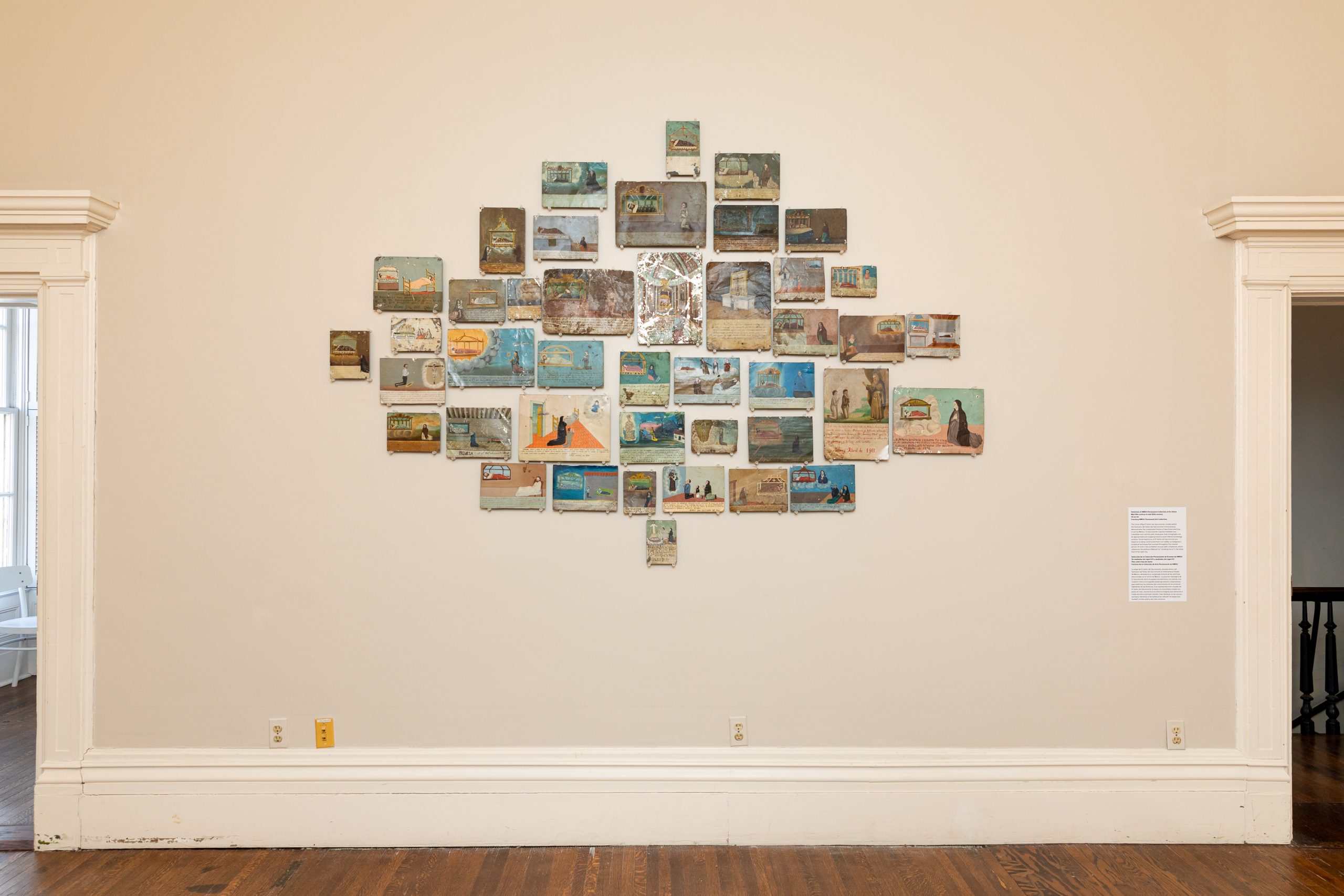 A grid of small paintings arranged in a loose cluster on a beige gallery wall, each depicting various scenes. The display is framed by two doorways with white trim and a polished wooden floor below.