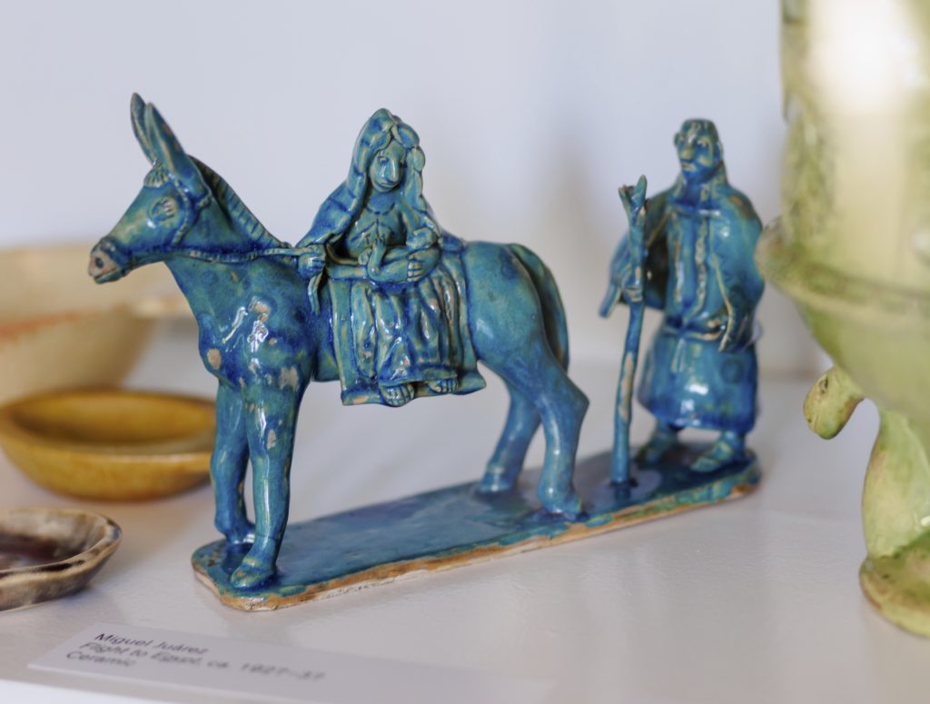 A blue ceramic figurine depicting a woman riding a donkey with a cloaked figure walking beside her, both on a rectangular base, displayed on a white shelf with other pottery items nearby.
