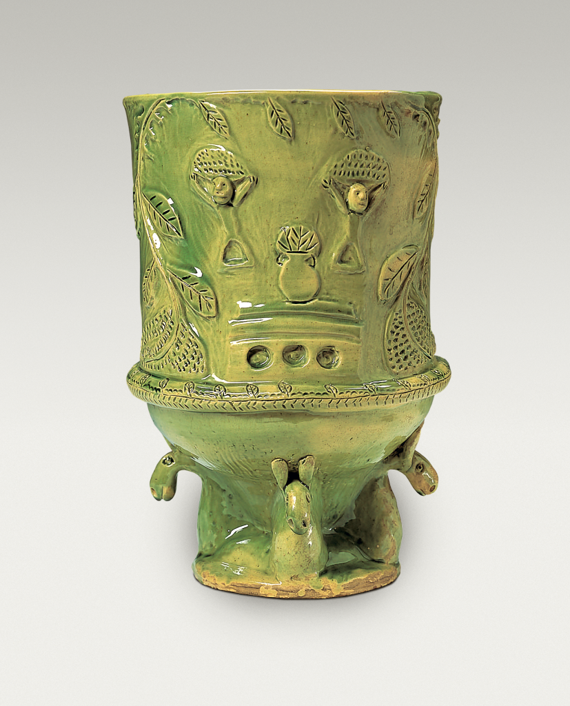 A green-glazed ceramic vessel with carved tree, bird, and flower motifs around the top and animal-shaped supports at the base, set against a plain background.