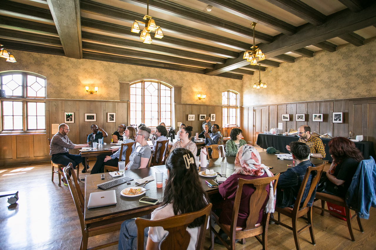 People sit around tables in a spacious, well-lit room with wooden beams and large windows, listening to a speaker at the front. Laptops, drinks, and papers are on the tables, and framed photos decorate the walls.