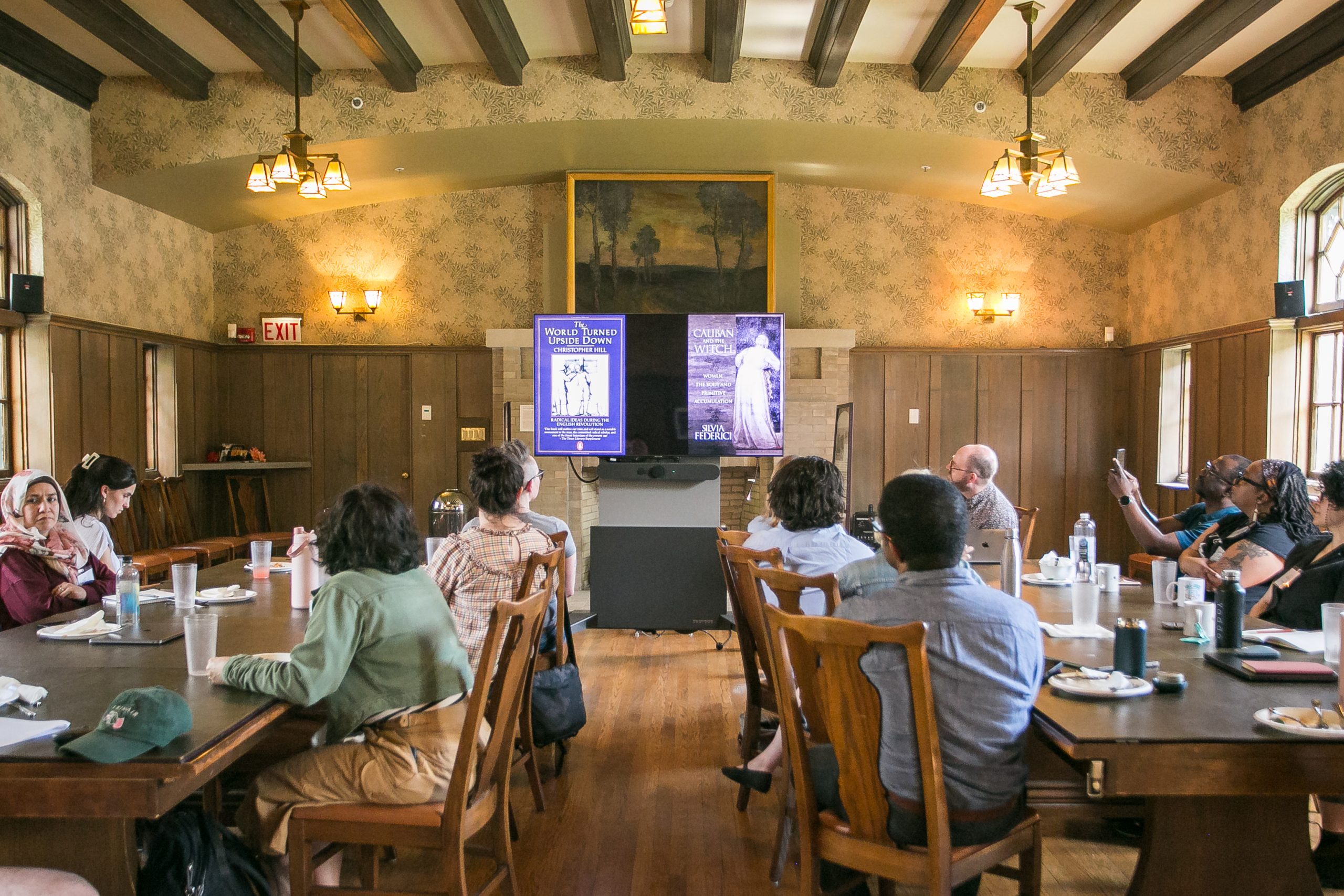 A group of people sit around tables in a wood-paneled room, watching a presentation displayed on two screens at the front. The room has large windows, high ceilings with beams, and food and drinks on the tables.