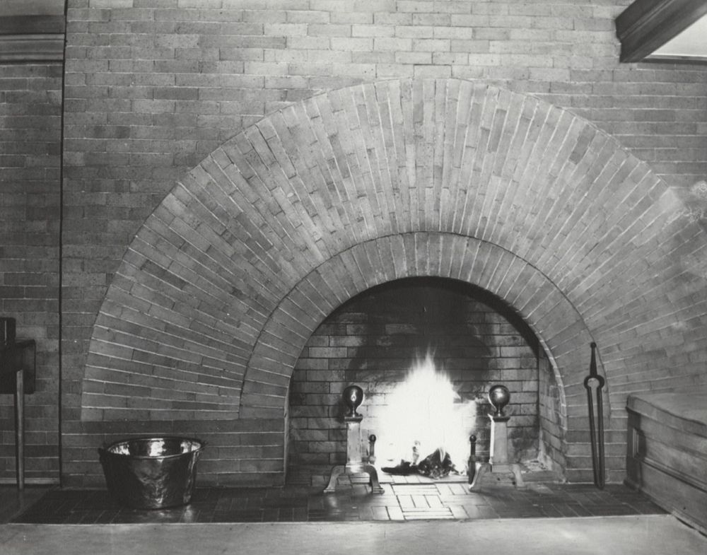 A brick fireplace with a large arched opening, a lit fire inside, two andirons, a metal bucket on the left, and a poker on the right. The bricks are arranged in a radiating pattern above the arch.