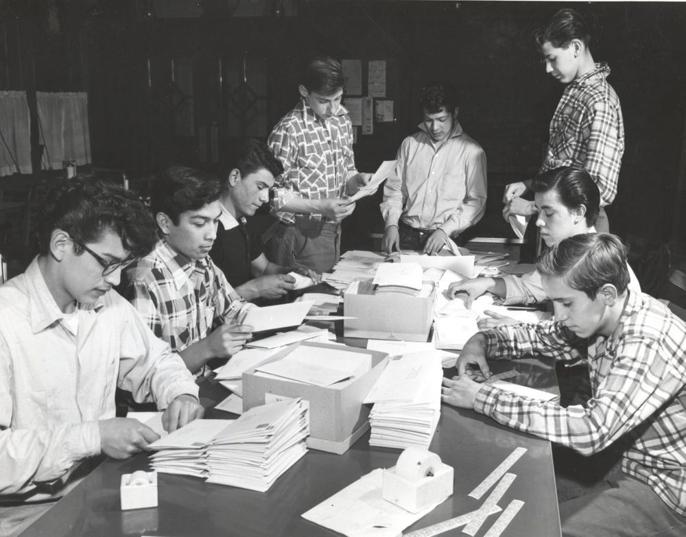 A group of young men sit and stand around a table sorting and assembling stacks of papers in a classroom or workshop setting, focused on their work.