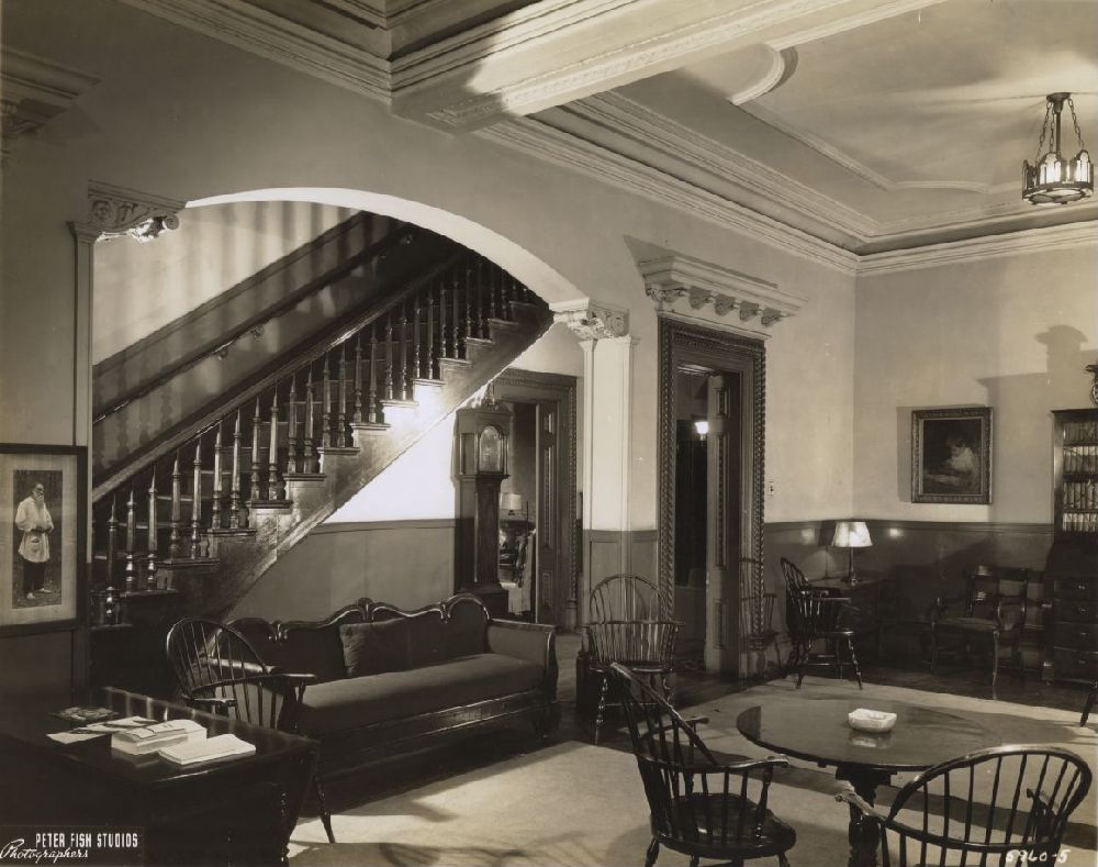 A vintage living room with wooden furniture, including chairs and a sofa, a round table, and a prominent staircase under an arched entryway. Framed pictures, lamps, and decorative molding adorn the space.