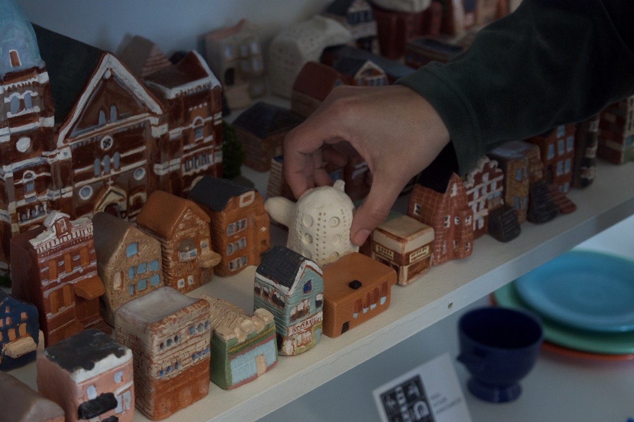 A hand picks up a small, white ceramic house from a shelf filled with miniature, colorful ceramic buildings and houses. Other pottery items are visible in the background.