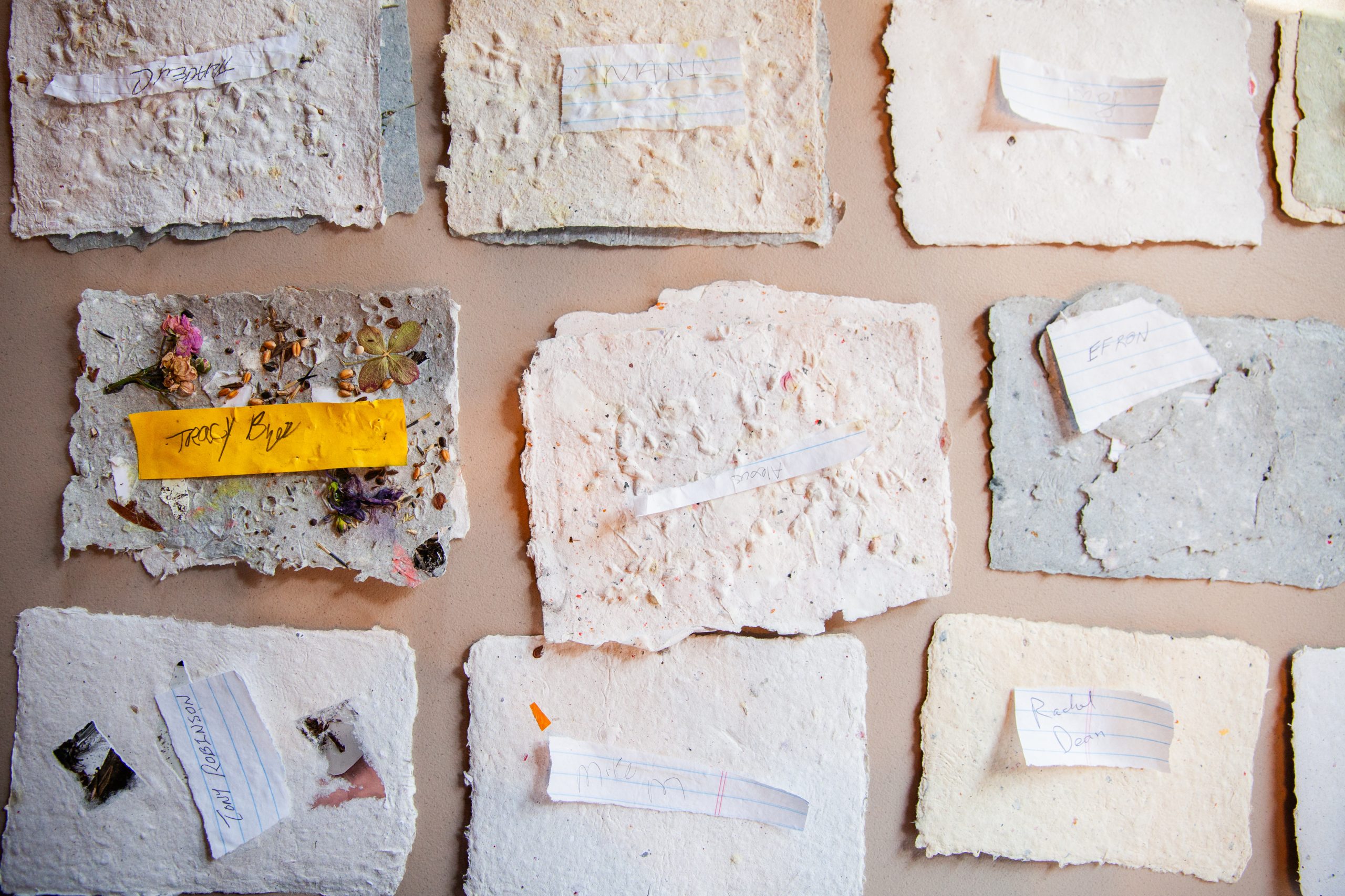 Several pieces of handmade paper, some with embedded flowers or leaves, are displayed on a flat surface. Each sheet has a small strip of paper with handwritten text placed or attached on top.