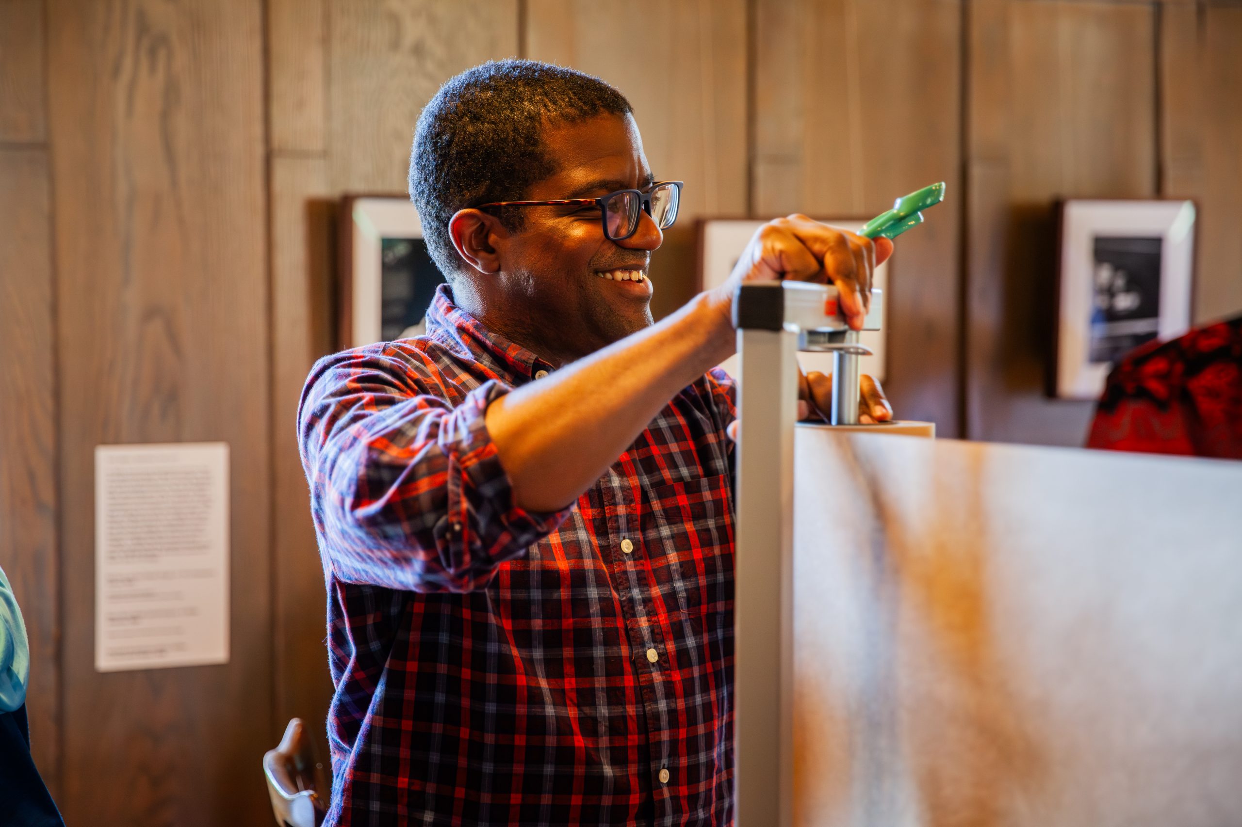 A man wearing glasses and a red plaid shirt smiles while assembling or adjusting a metal stand in a warmly lit room with wood-paneled walls and framed pictures.
