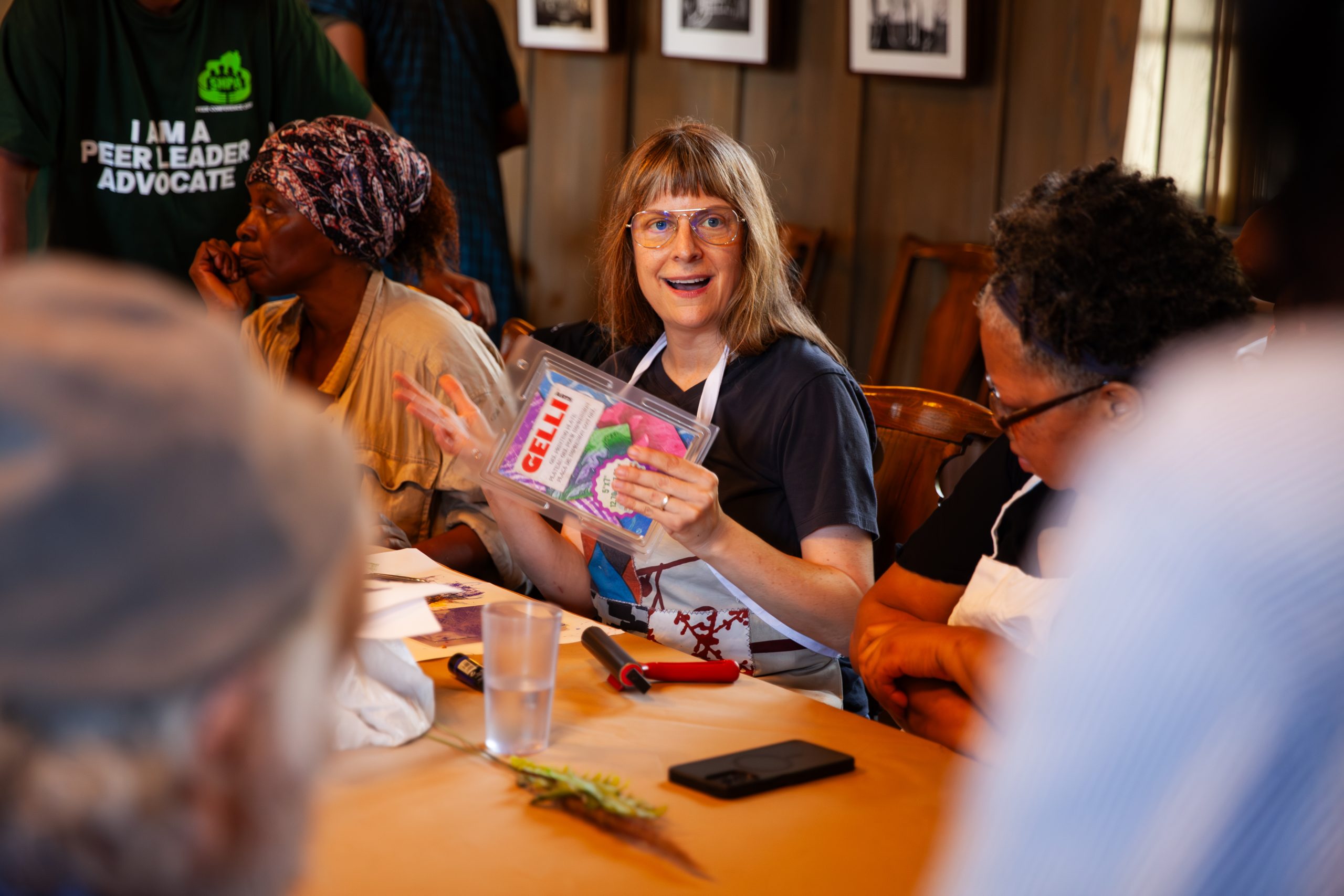 A woman sits at a table holding up a seed packet and smiling, surrounded by other people engaged in conversation during a community gathering or workshop.