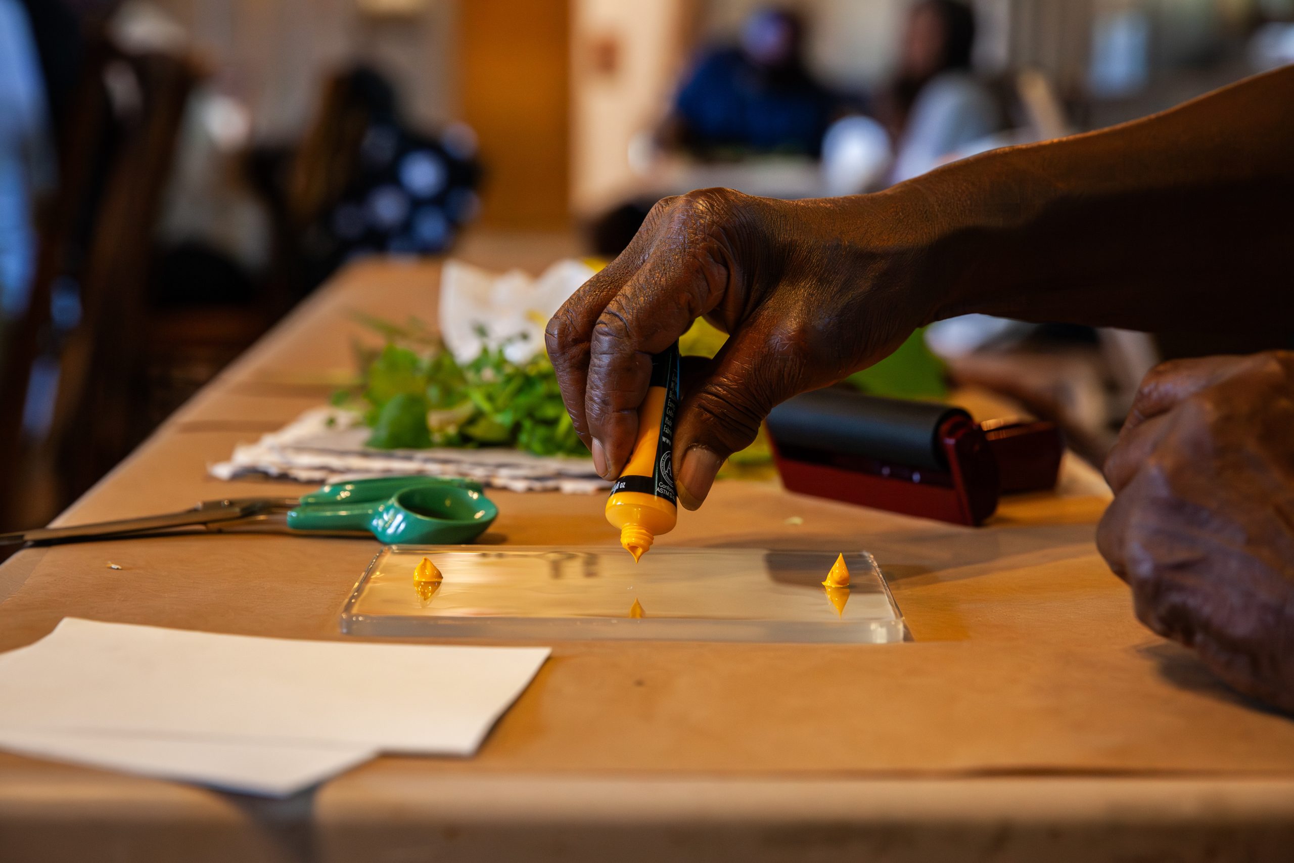 A hand squeezes yellow paint from a tube onto a flat surface, with scissors, green leaves, and other objects on a table in the background. The setting appears to be a creative or craft workspace.