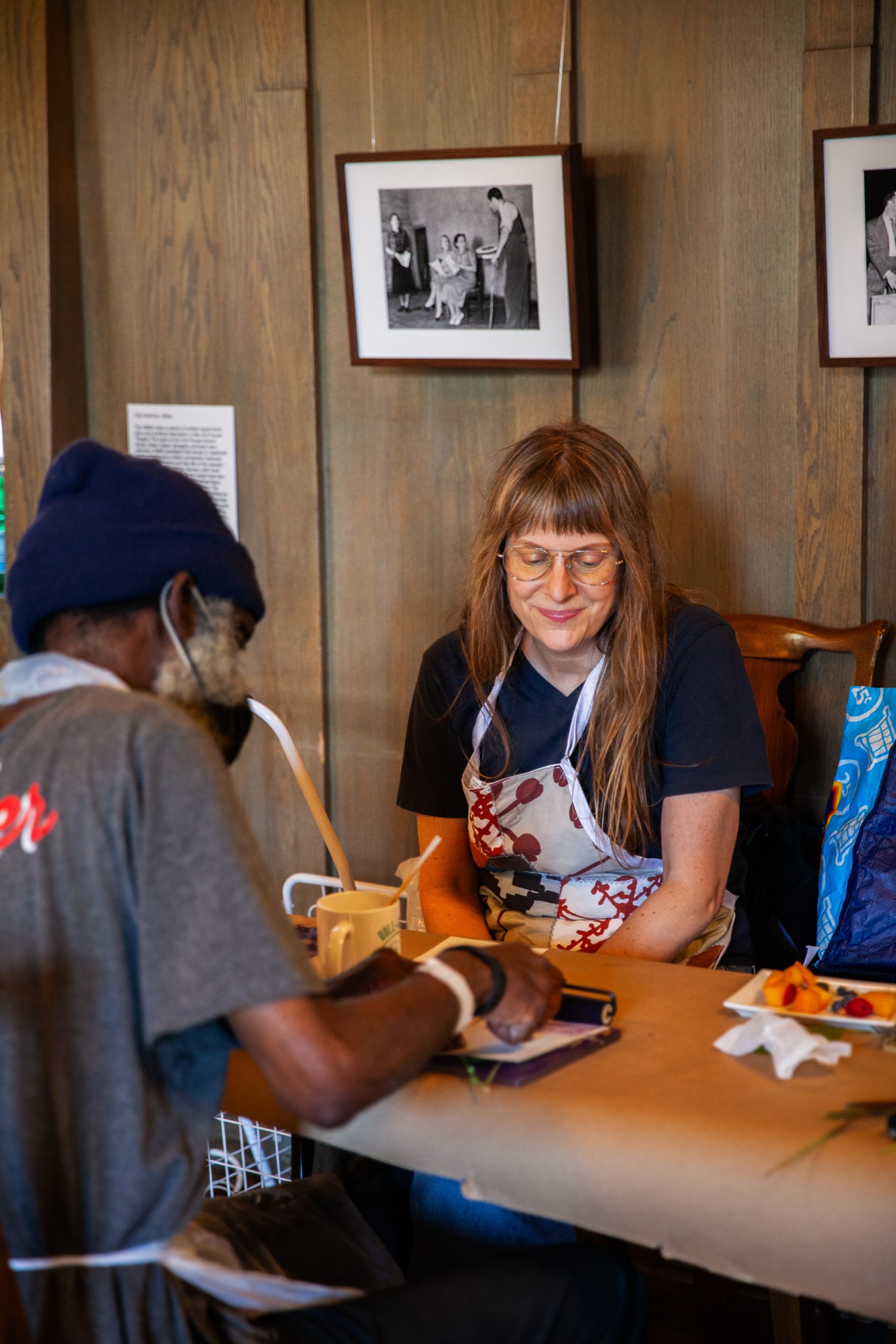 A woman with long hair and glasses smiles while sitting at a table across from a man who is writing or drawing. They are in a cozy, wood-paneled room with framed photos on the wall behind them.